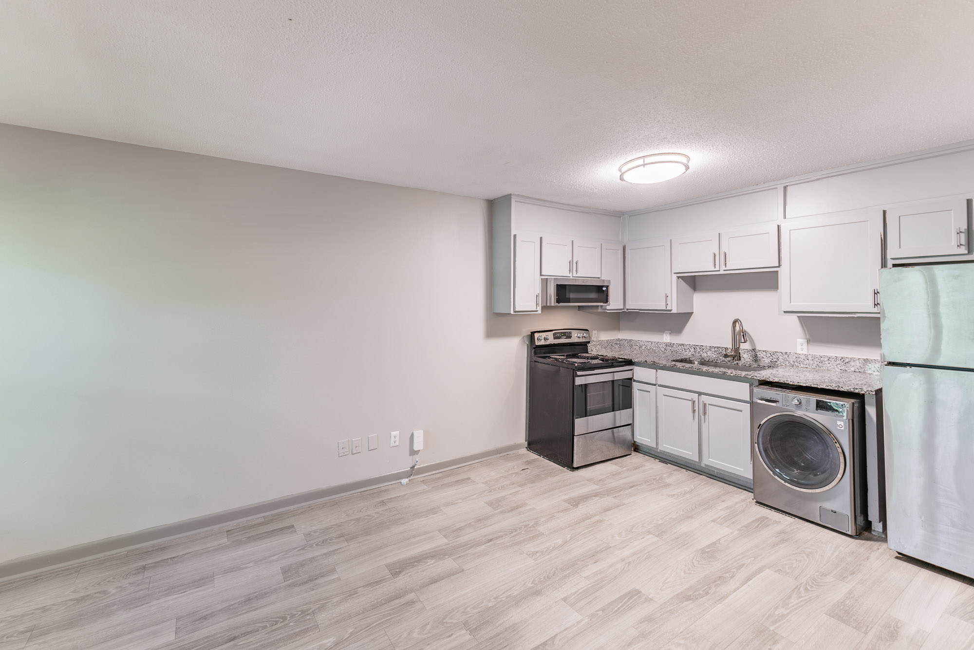 Modern, well-lit kitchen featuring gray cabinets, stainless steel appliances, a granite countertop, and a washer/dryer. The floor is light-colored wood-like laminate, and the walls are a soft gray. Overall, the space is clean and contemporary, perfect for a cozy home atmosphere.