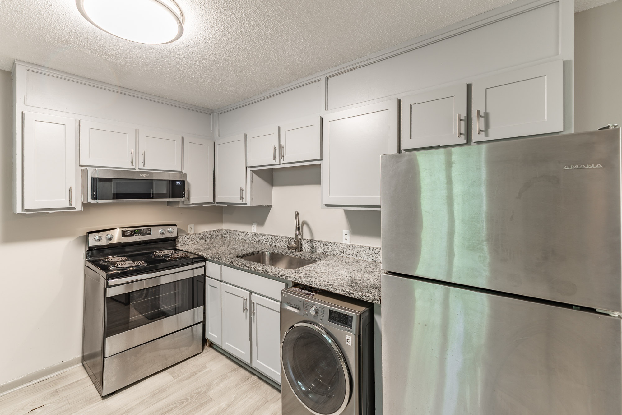 Modern kitchen featuring gray cabinets, stainless steel appliances including a stove, microwave, refrigerator, and a stacked washer and dryer. The countertop is made of granite, and the space is well-lit with natural light. The flooring is light-colored, enhancing the overall clean and contemporary look.