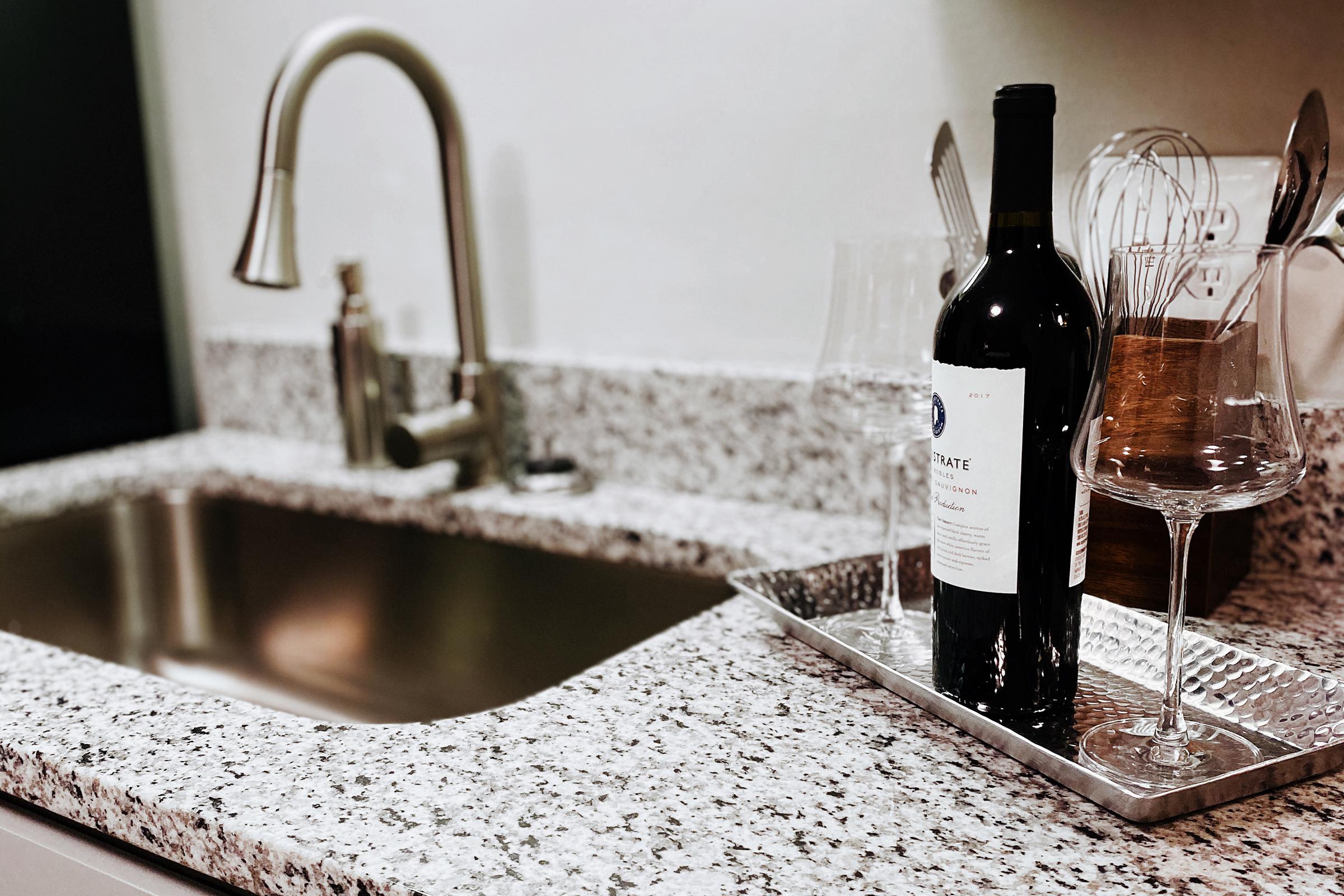 A modern kitchen sink made of stainless steel, surrounded by a granite countertop. A bottle of red wine stands on a silver tray alongside two elegant wine glasses, with kitchen utensils in the background. The setting is well-lit and organized, creating an inviting atmosphere.