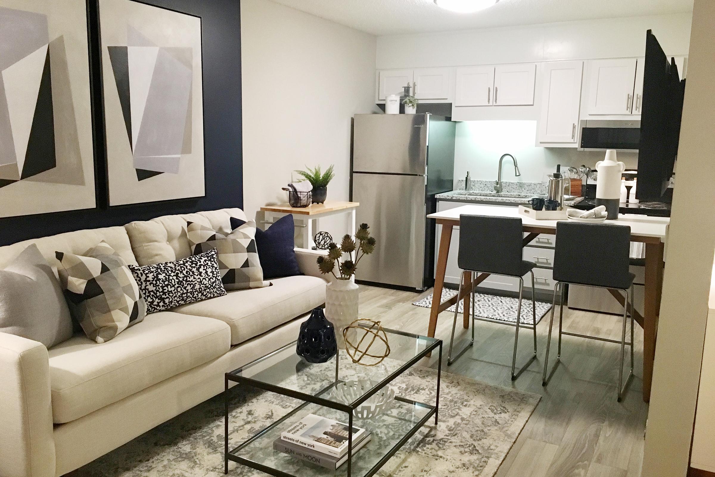 A modern living space featuring a comfortable cream-colored sofa with patterned cushions, a coffee table with decorative items, and two barstools at a kitchen island. The kitchen has white cabinetry and stainless steel appliances. Abstract artwork decorates the walls, and a plant adds a touch of greenery.