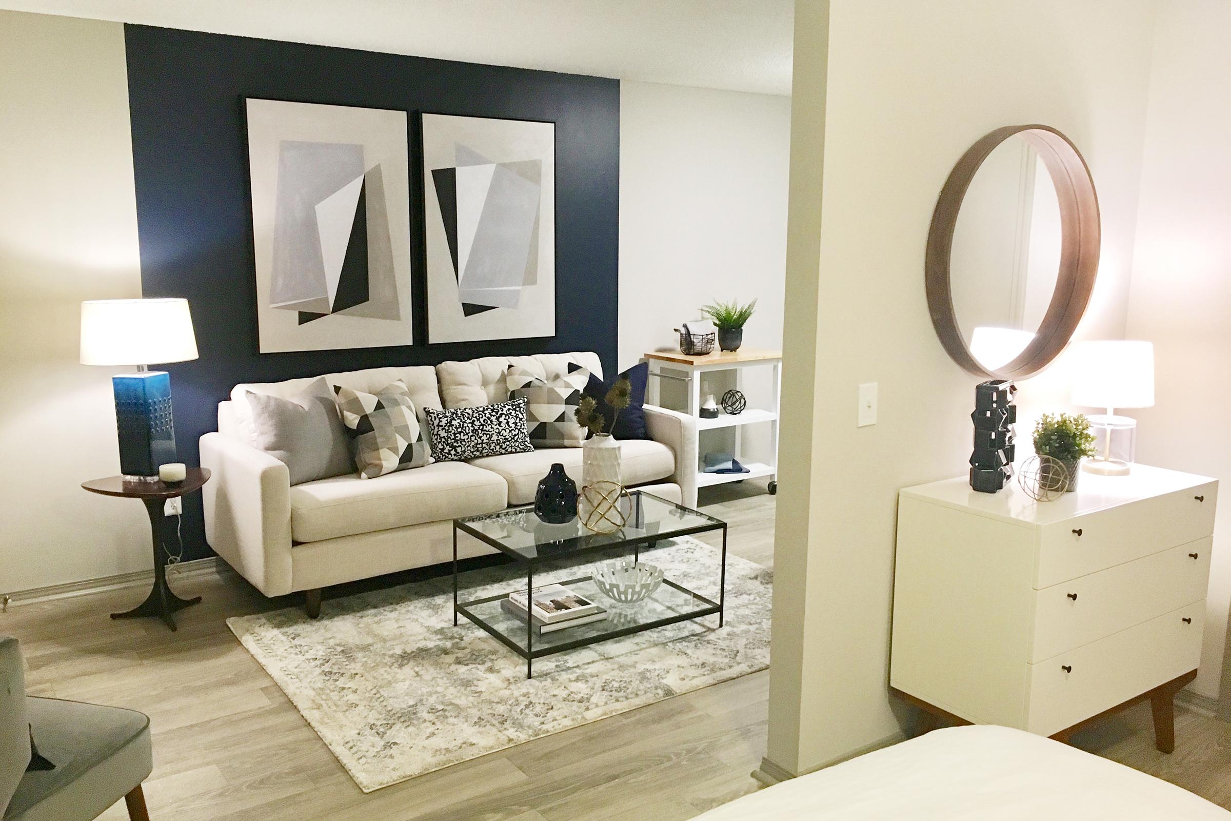 A modern living room featuring a white sofa with decorative pillows, a glass coffee table, and a patterned area rug. There are two abstract art pieces on the wall, a round mirror, and lamps providing soft lighting. A stylish side table and a white dresser complete the cozy and contemporary atmosphere.