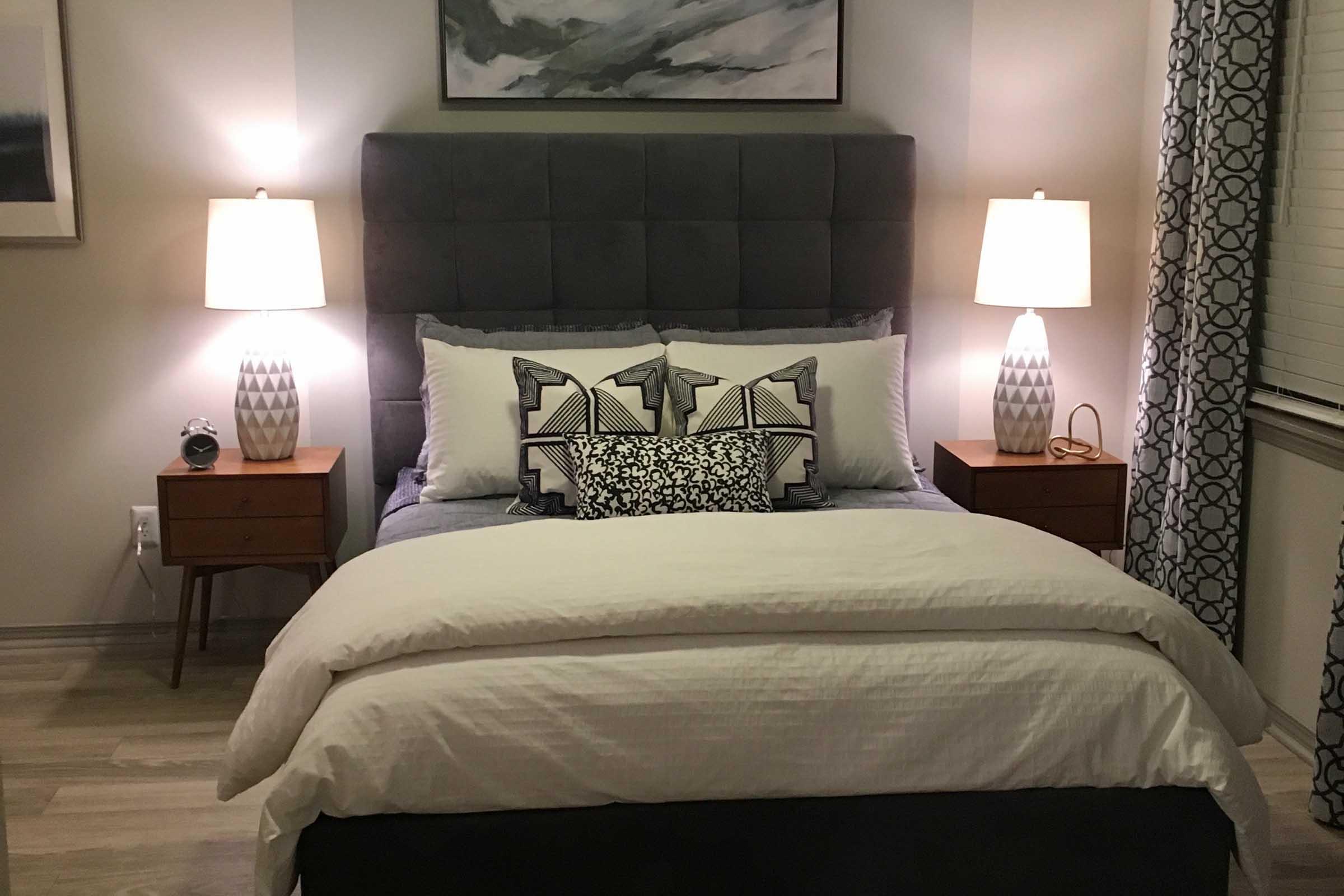 A cozy bedroom featuring a dark gray upholstered headboard and white bedding. Two stylish bedside lamps illuminate wooden nightstands on either side of the bed. Decorative pillows are arranged at the head of the bed. A piece of abstract art hangs above the headboard, and patterned curtains frame a window.