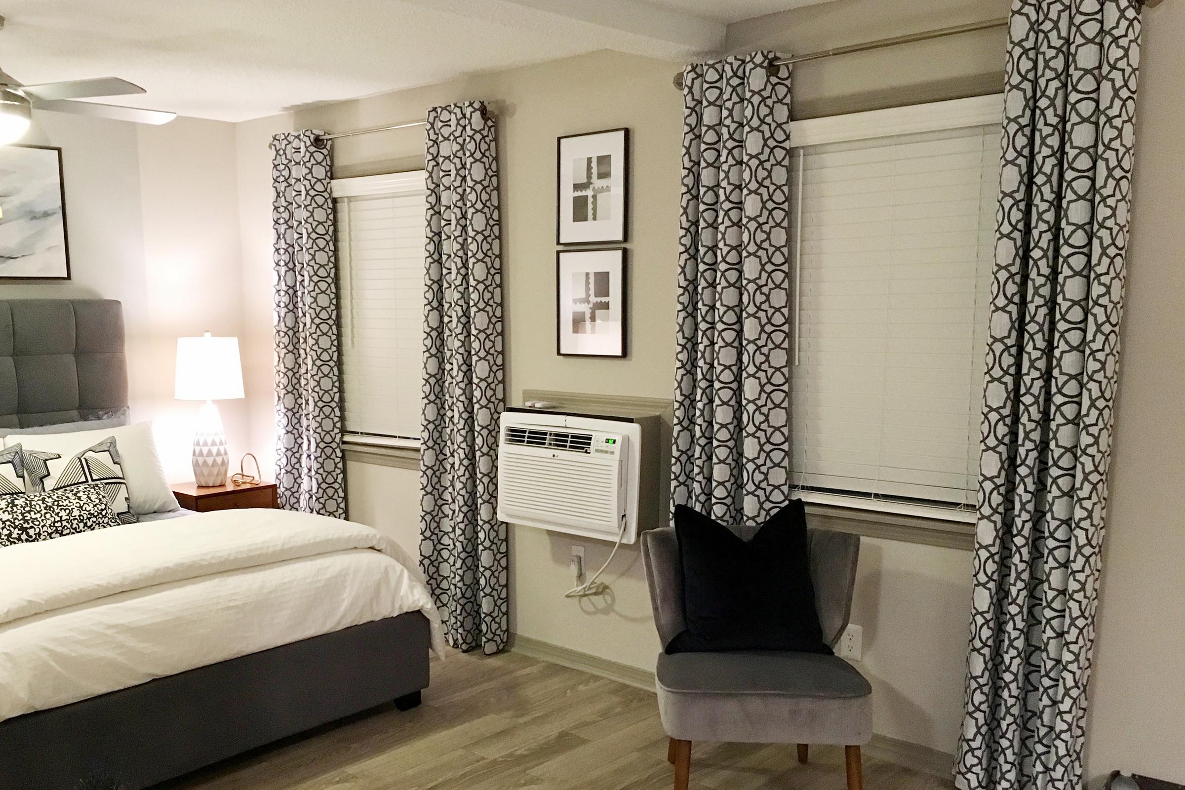 A cozy bedroom featuring a bed with decorative pillows, gray bedding, and a stylish headboard. Two windows are adorned with patterned gray curtains, and there's an air conditioning unit below one of the windows. A modern accent chair sits in the corner, and framed artwork is displayed on the walls.