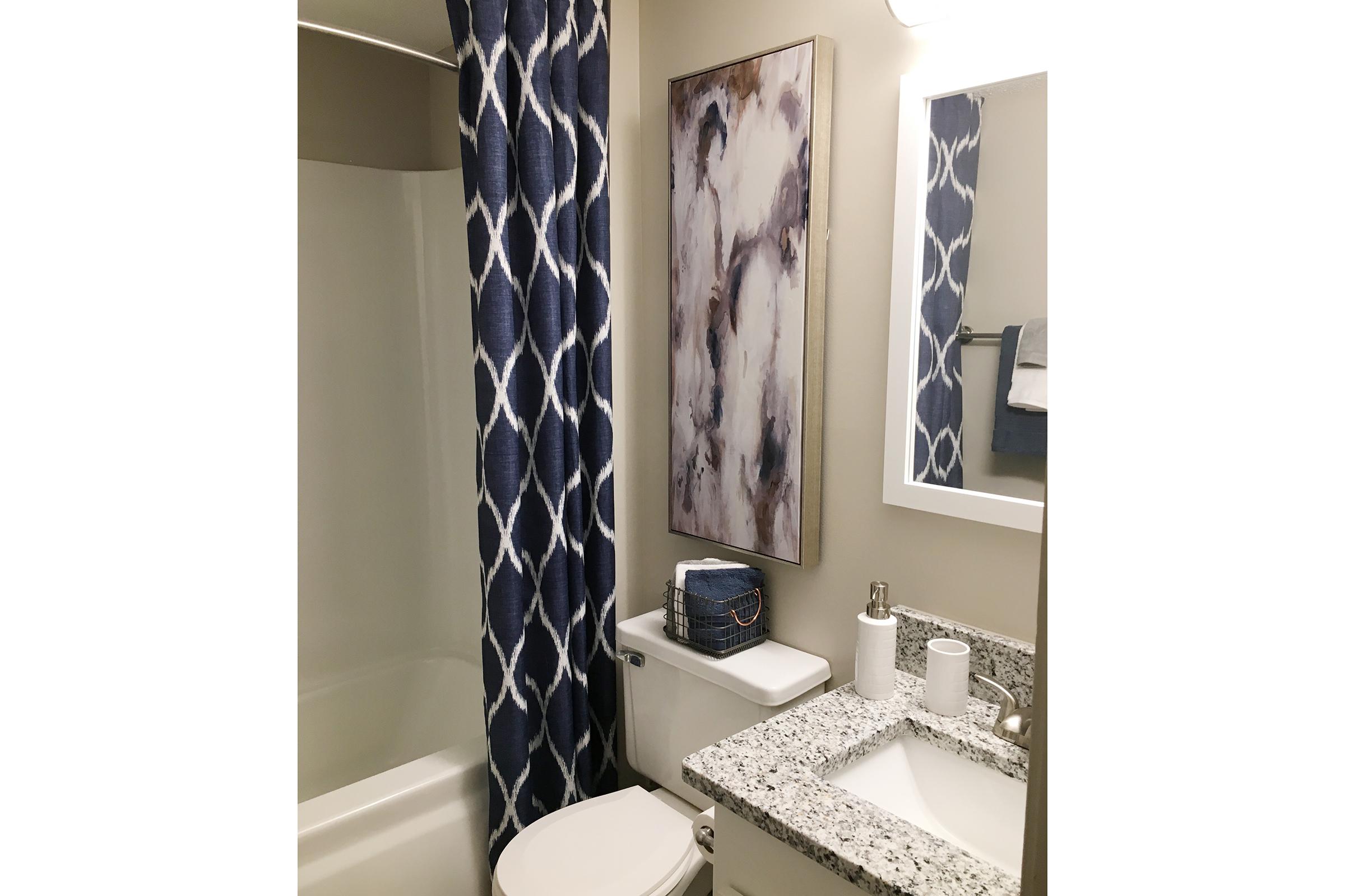 A modern bathroom featuring a white bathtub with a blue and white patterned shower curtain, a granite countertop with a small gray storage box, a mirror, and wall art. The bathroom is well-lit and organized, showcasing a clean and stylish design.