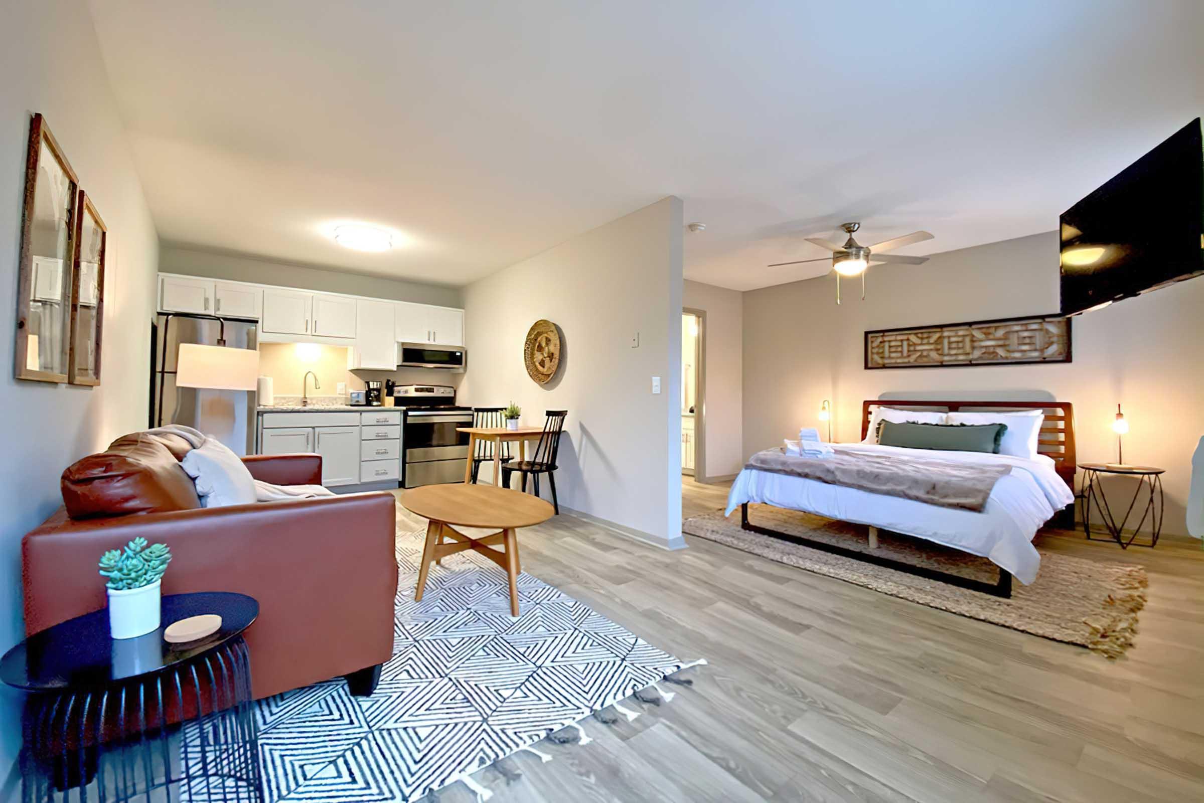 A cozy studio apartment featuring a brown leather sofa, a round wooden coffee table, and a queen-sized bed with white linens. The kitchen area has modern appliances and a dining table with chairs. A large wall-mounted TV and decorative elements like a wall hanging and a plant complete the space.