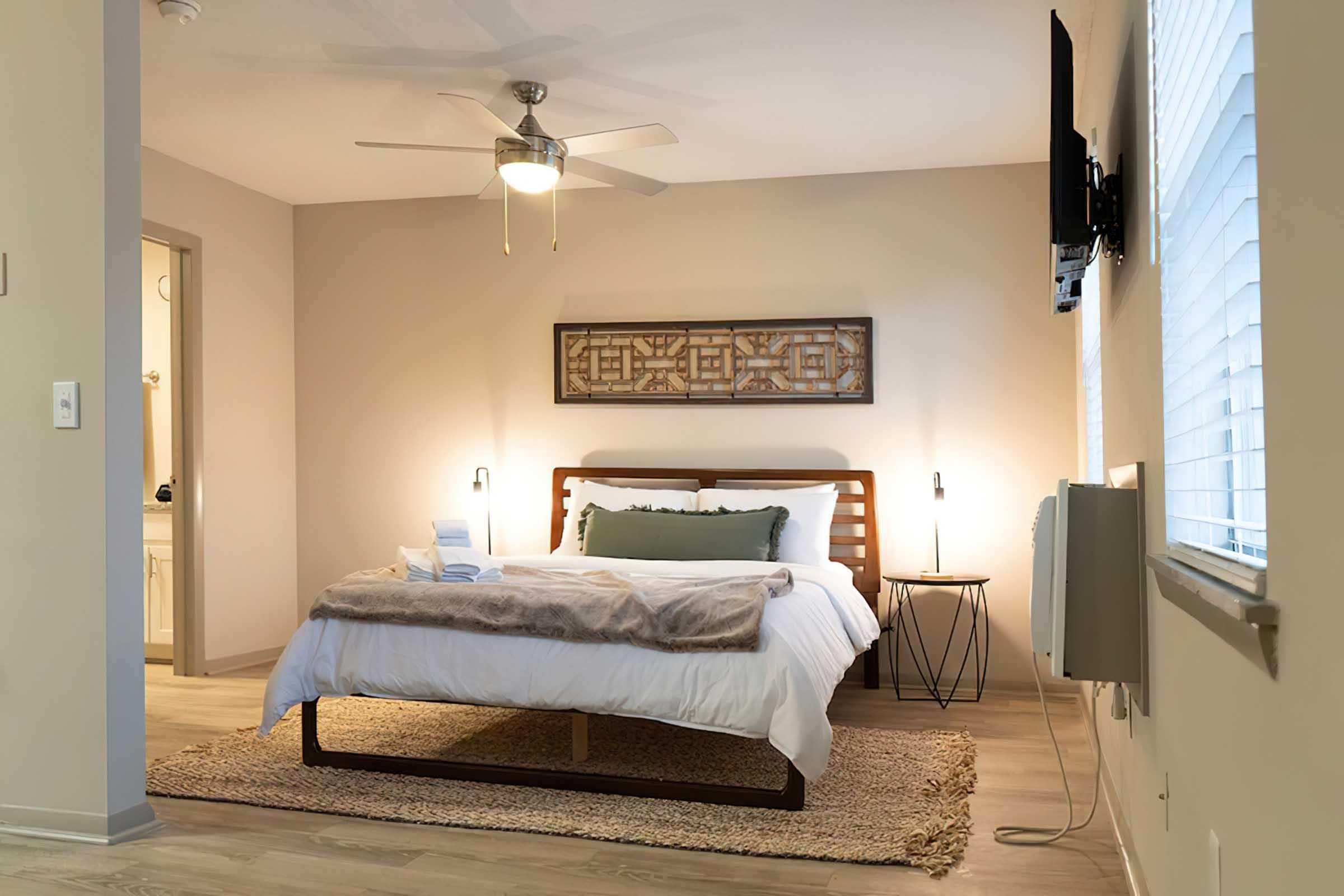 Cozy bedroom featuring a large bed with white linens and a gray throw, wooden accents, and a stylish headboard. A ceiling fan and bedside lamps enhance the ambiance. A textured area rug adds warmth, and a flat-screen TV is mounted on the wall. Natural light filters through window blinds, creating a serene atmosphere.