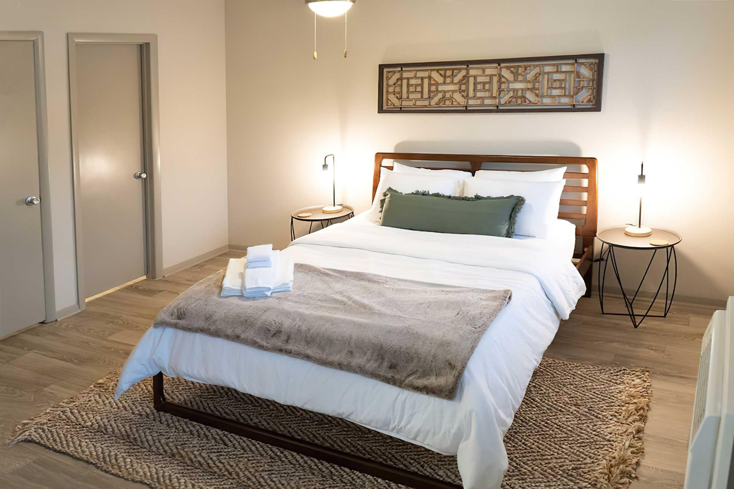 A cozy bedroom featuring a wooden bed with white bedding and a green accent pillow. Two nightstands with lamps are on either side, and a textured throw blanket is draped over the foot of the bed. Light-colored walls and a woven rug add warmth to the space, with a subtle decorative wall hanging above the bed.
