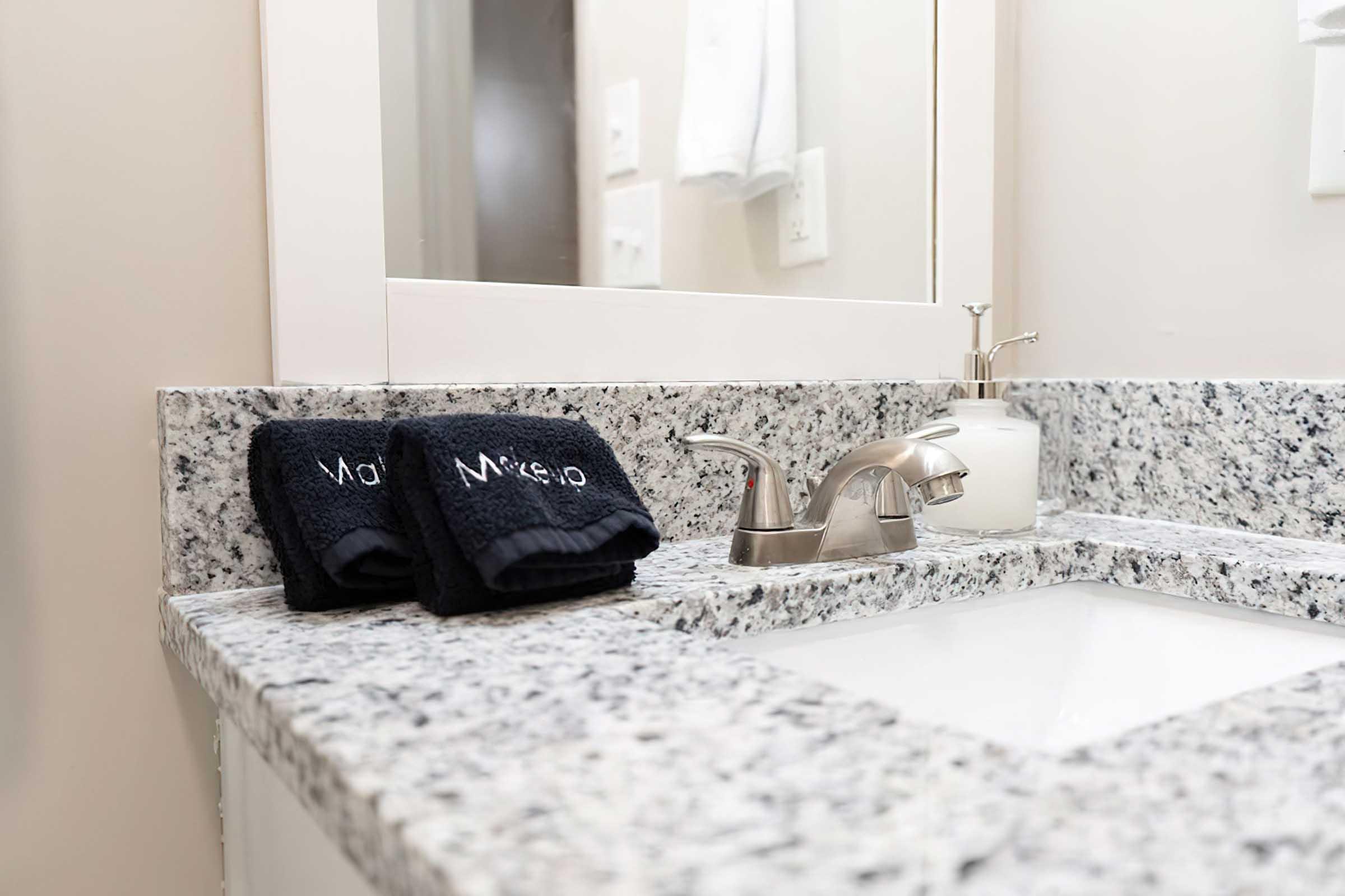 A modern bathroom counter featuring a white sink, a stylish faucet, two neatly folded dark towels with the word "Mako" embroidered on them, and a small, elegant soap dispenser. The counter is made of speckled granite, complemented by a light background and a mirror reflecting the overall clean aesthetic.