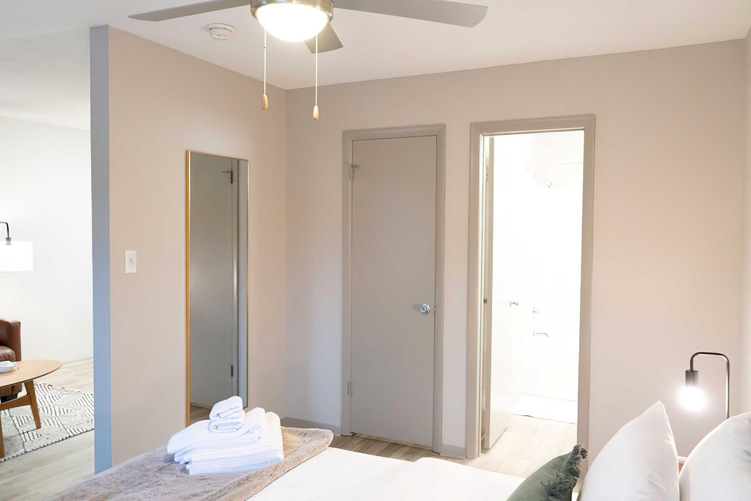 A well-lit bedroom featuring a bed with neatly arranged towels, a ceiling fan, a mirror, and a door leading to a bathroom. The walls are painted in a soft, neutral color, and there is a small light fixture beside the bed. The overall decor is modern and minimalistic.