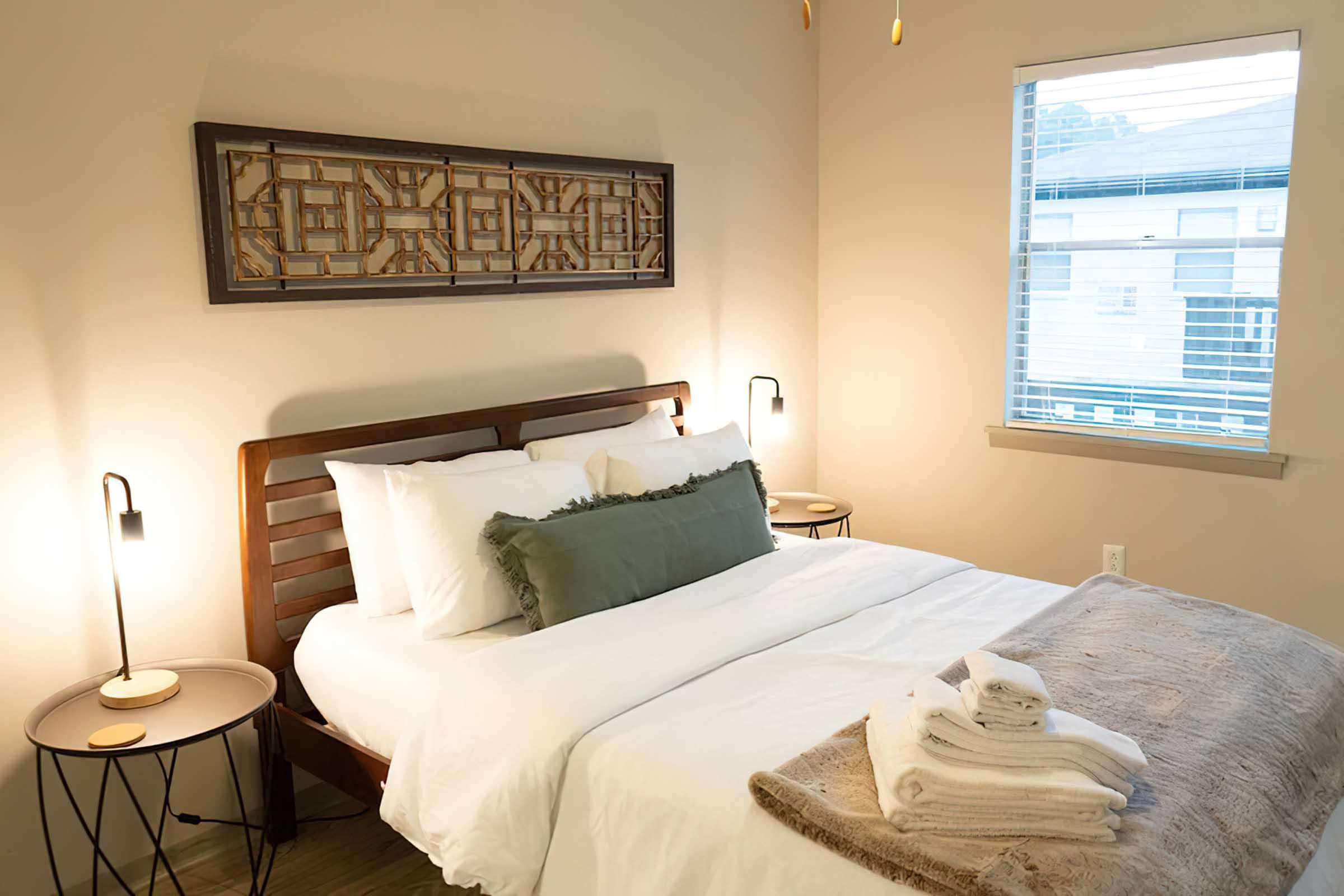 Cozy bedroom with a wooden bed frame, white bedding, and decorative pillows. A light-colored wall holds a framed artwork above the bed, and two bedside lamps provide soft lighting. A small round table next to the bed features folded towels. Natural light filters through a window with white blinds.