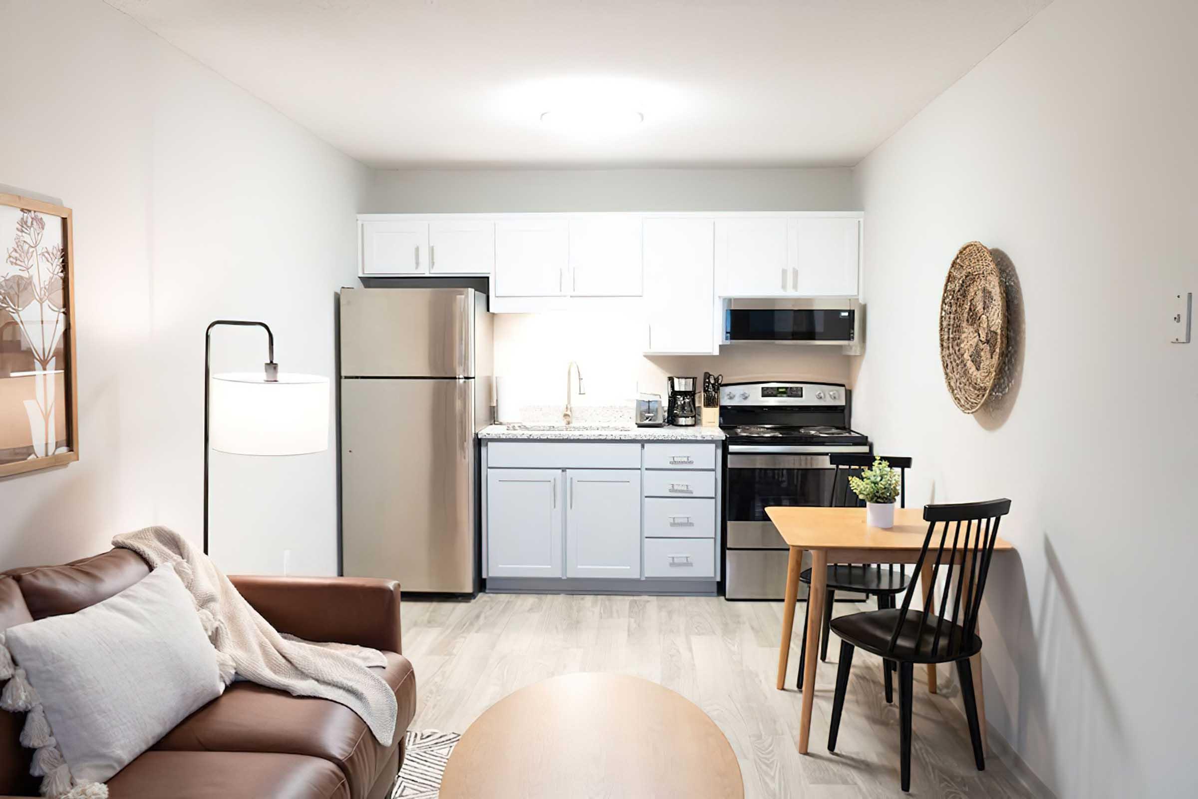 A modern kitchen and living area featuring a stainless steel refrigerator, stove, and microwave. The kitchen has white cabinetry and a light countertop. A cozy brown sofa is in the foreground, paired with a wooden coffee table. A small dining table with a black chair is visible, enhancing the inviting atmosphere.
