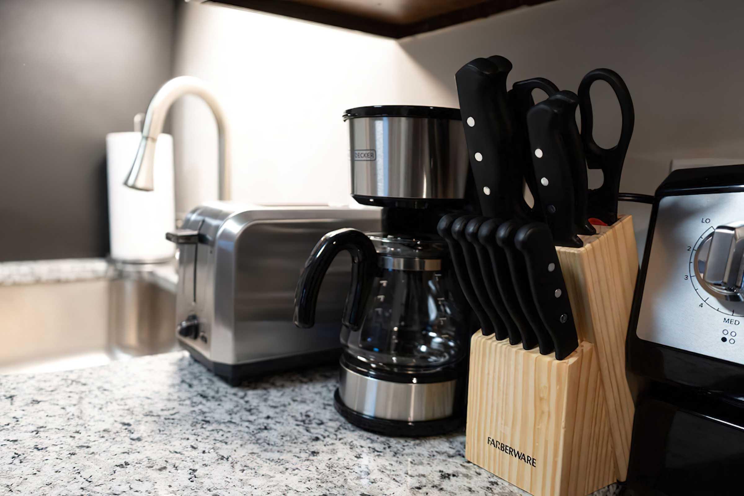A modern kitchen countertop featuring a coffee maker, a stainless steel toaster, a knife block filled with black-handled knives, and a sleek mixer. The countertop is made of speckled gray granite, and there is a sink in the background with a faucet. Soft lighting adds a warm ambiance to the space.