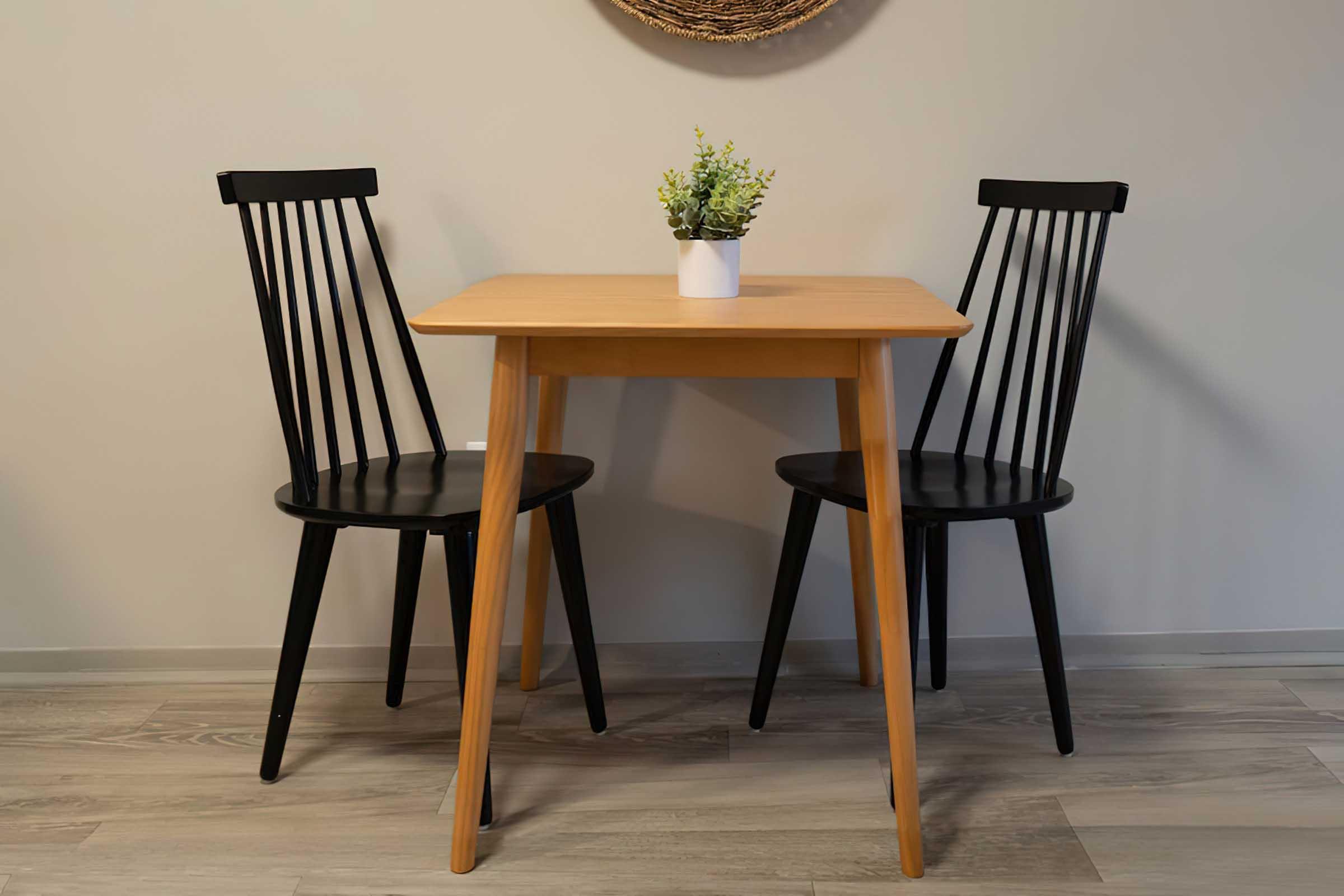 A wooden table with two black chairs on either side. The table has a small white vase with green foliage on top. The setting has a neutral wall and a decorative circular wall hanging in the background. The floor is light-colored wood.