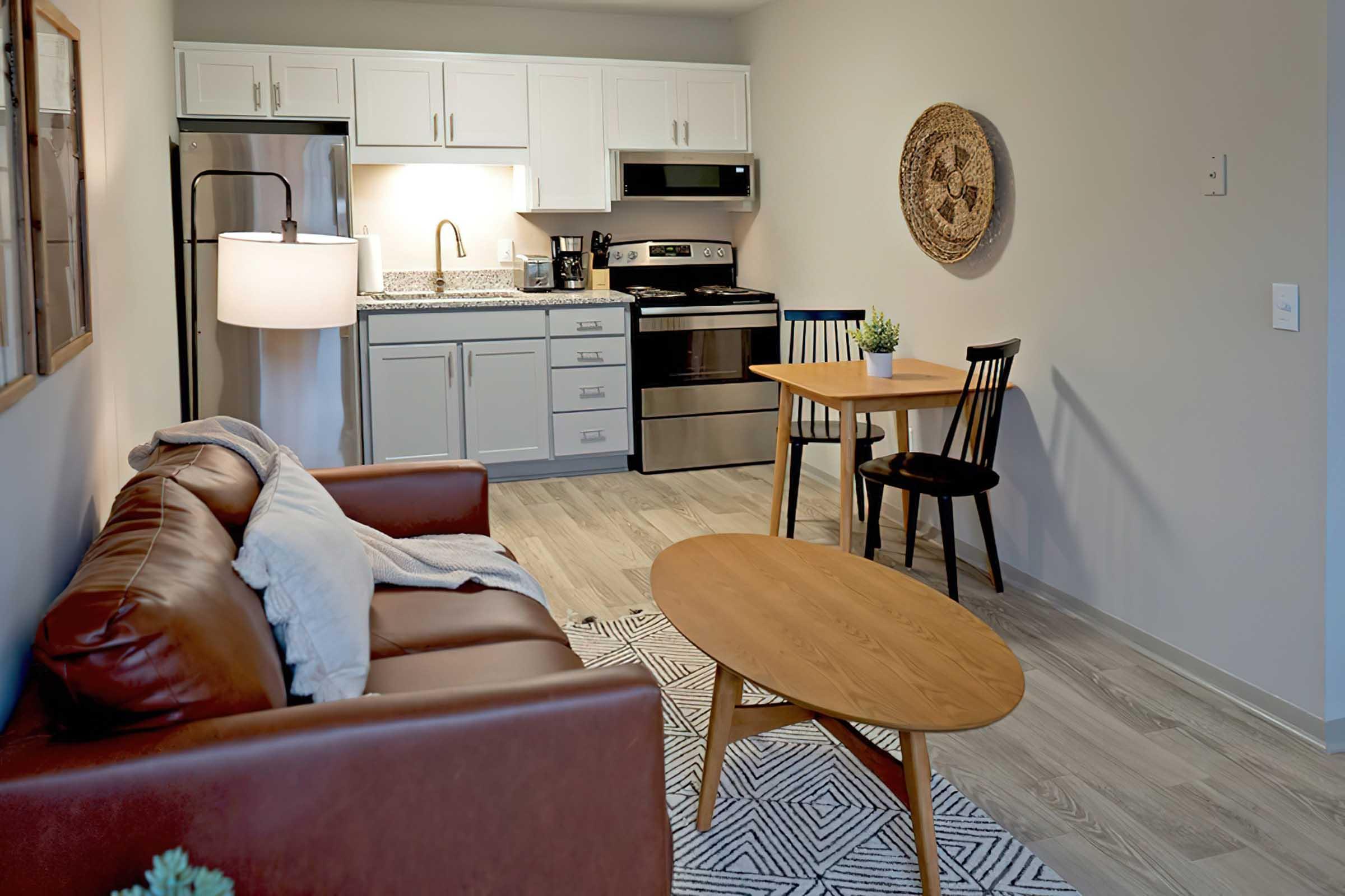 A cozy kitchen and dining area featuring a brown leather sofa, a round wooden coffee table, and two black chairs. The kitchen includes a fridge, stove, and cabinetry with a modern design. Natural light fills the space, highlighting a decorative wall piece and a plant on the table.