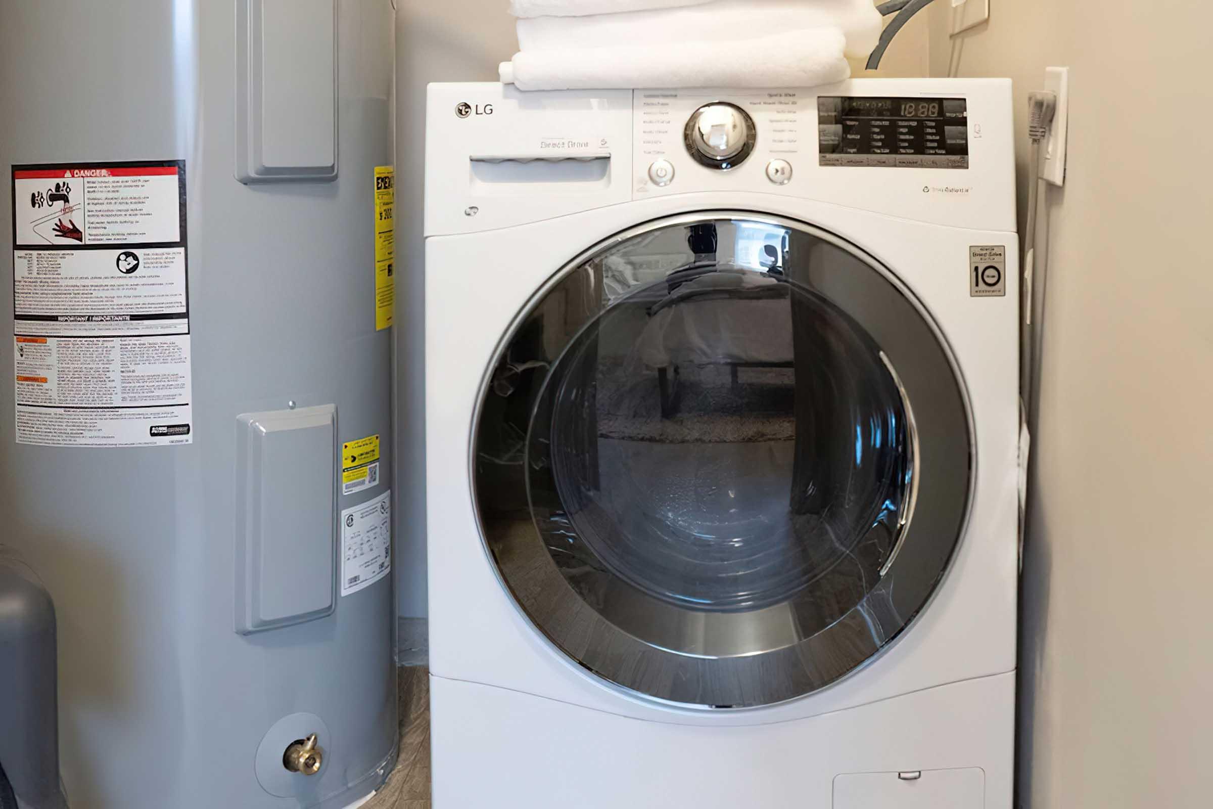 A modern LG front-loading washing machine situated next to a water heater in a laundry area. The washing machine features a transparent door, control panel with various settings, and is stacked with neatly folded white towels. The surrounding walls are painted in neutral tones, enhancing the clean, organized look.