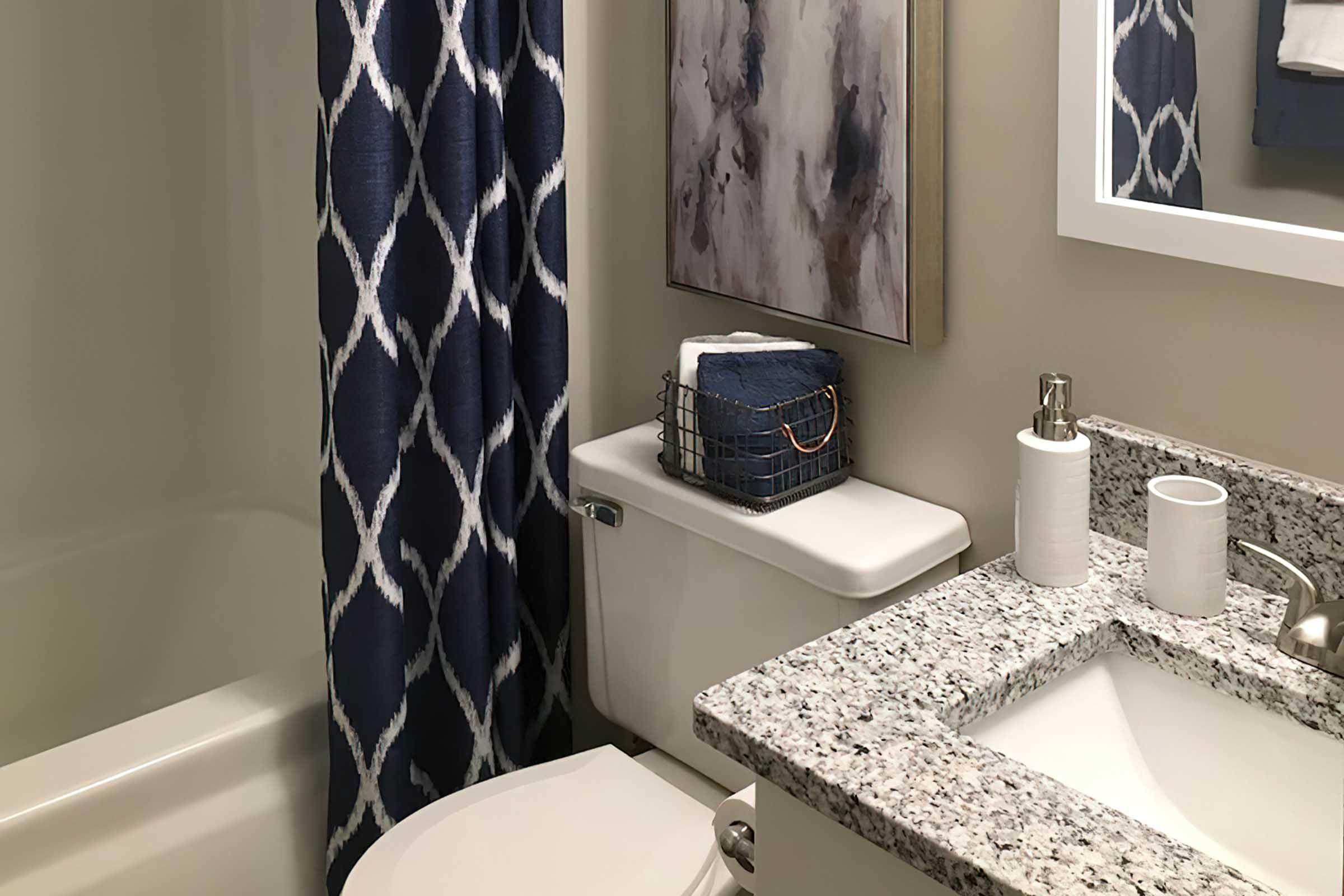 A small bathroom featuring a bathtub, toilet, and a granite countertop with a sink. The wall is adorned with a decorative mirror and a framed abstract artwork. A navy blue shower curtain with a geometric pattern adds color, and a basket on the counter holds toiletries.