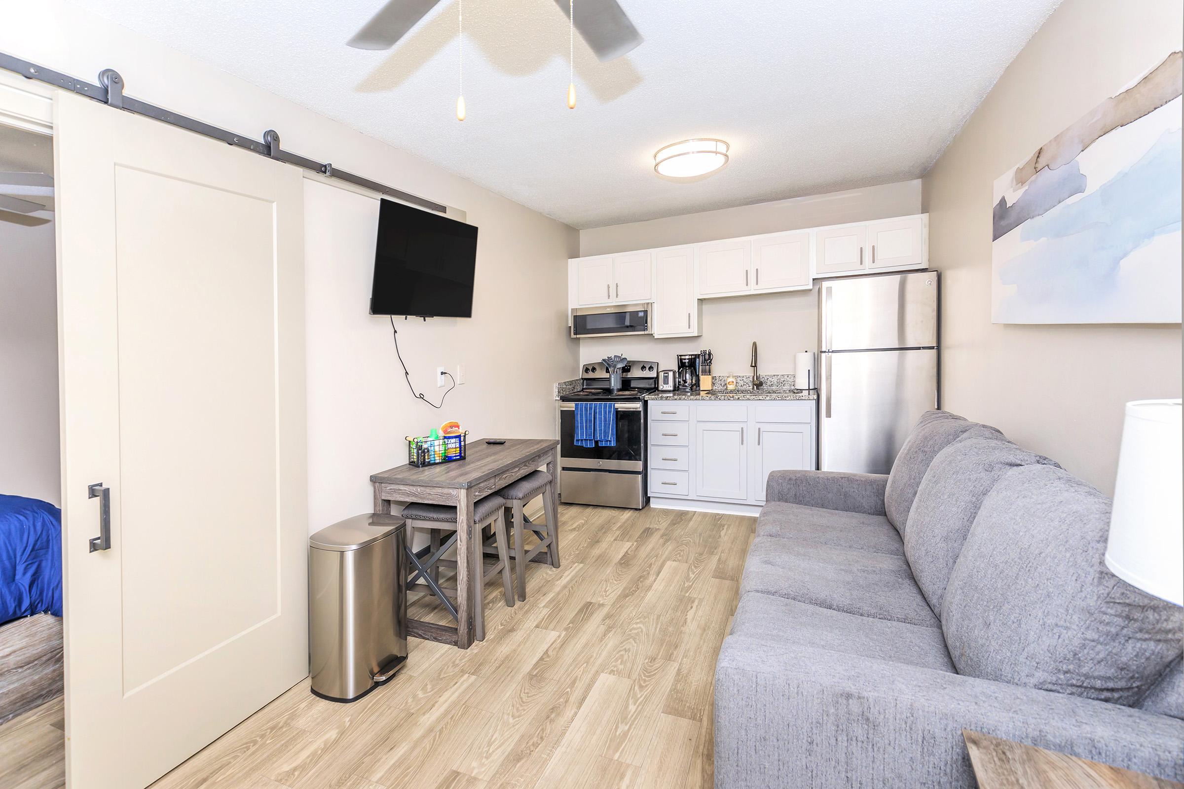 A modern living space featuring a gray sofa, a wooden dining table, and a flat-screen TV mounted on the wall. The kitchen area includes stainless steel appliances, including a fridge and stove, with light-colored cabinets. The flooring is a light wood, adding a warm feel to the room.