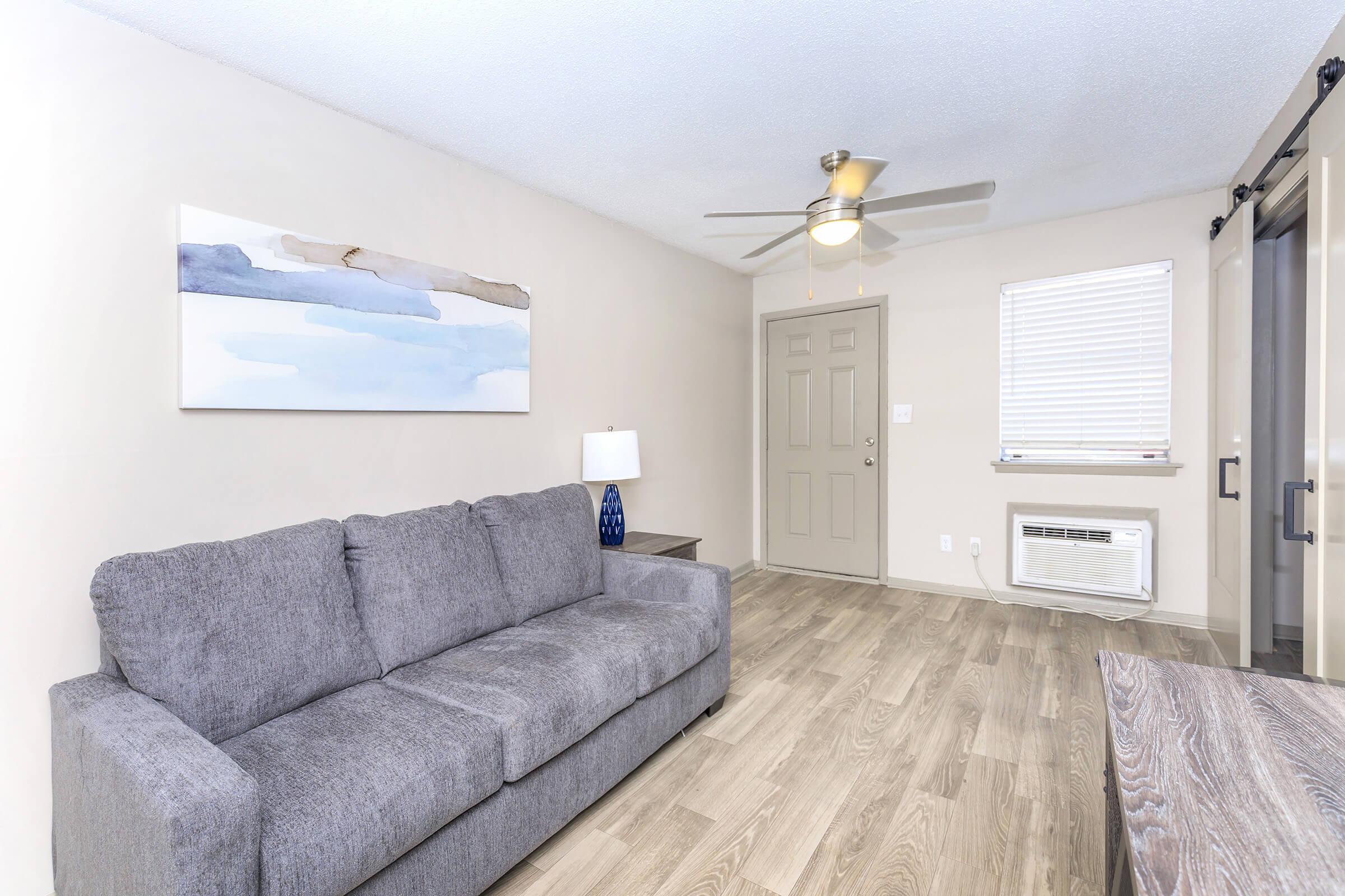 A modern living room featuring a gray sofa, a blue table lamp, and a decorative wall piece in shades of blue. There’s a door leading outside, a window with blinds, and an air conditioning unit. The flooring is light wood, creating a cozy atmosphere.