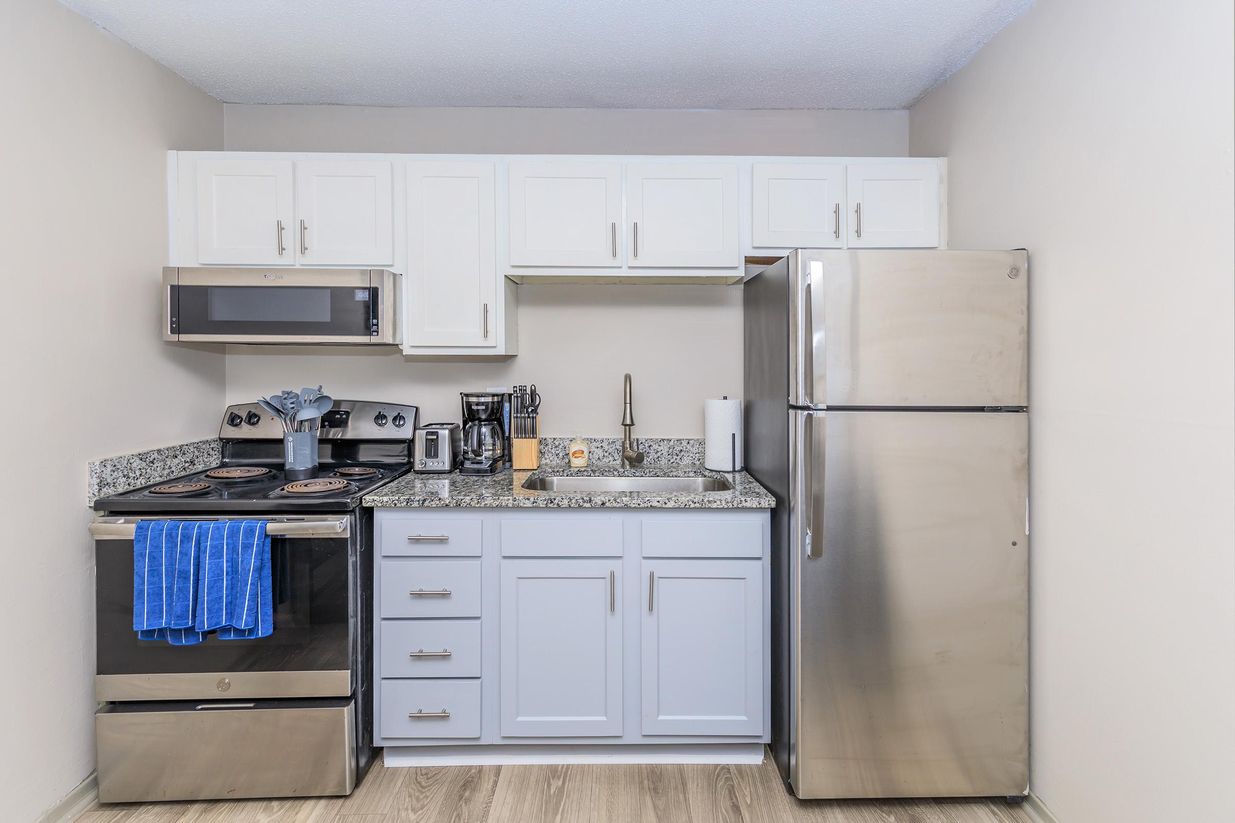 Compact kitchen featuring modern appliances, including a stainless steel refrigerator, a microwave, an oven with a stovetop, and a coffee maker. The cabinets above are white, contrasting with the dark countertop. A blue kitchen towel hangs from the oven, and the flooring is light-colored wood.