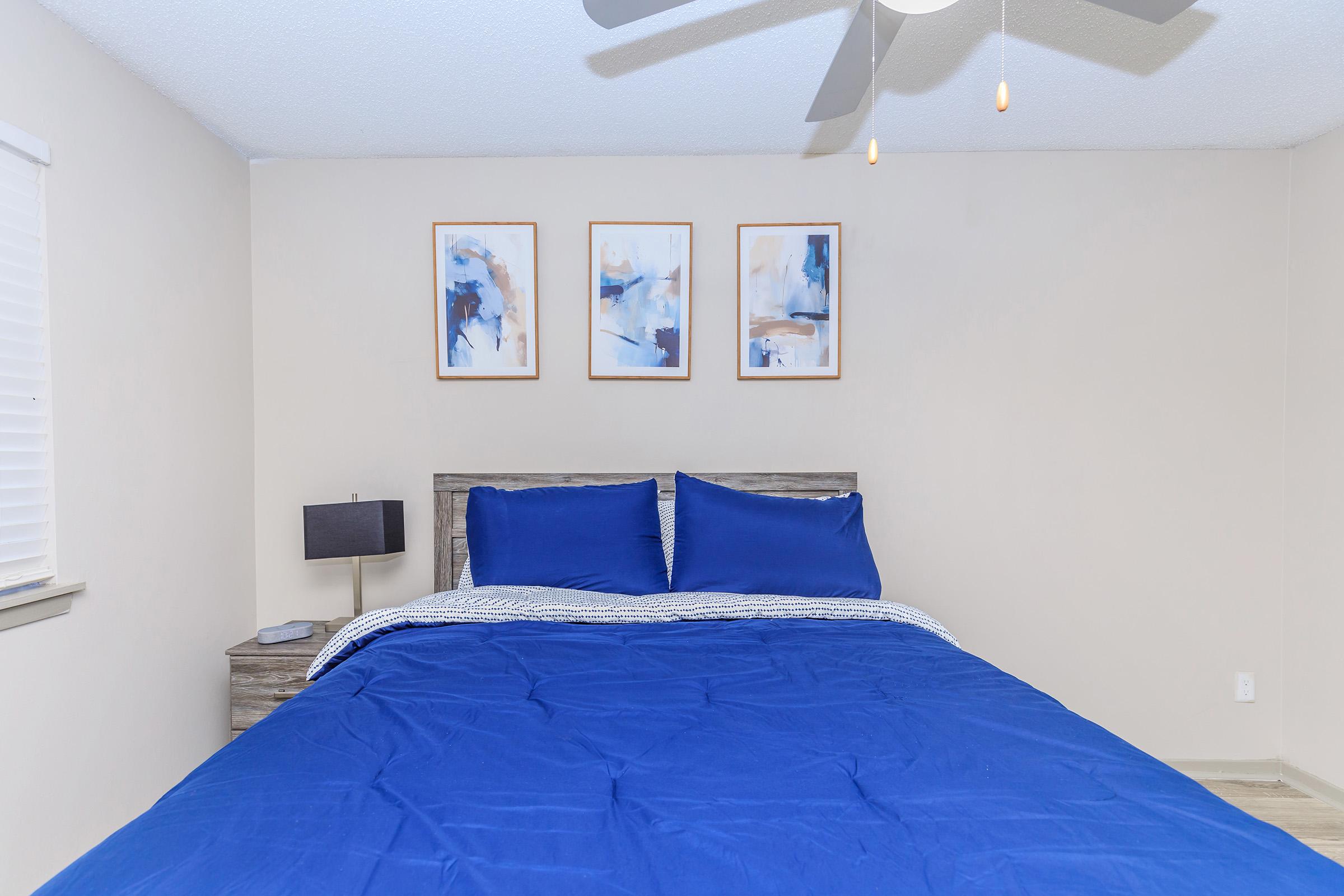 A cozy bedroom featuring a large blue bedspread on a wooden bed frame, two blue pillows, and a decorative blanket. On the wall, there are three framed abstract paintings in shades of blue. A small bedside lamp sits on a wooden nightstand, and there is good natural light from a window with white blinds.