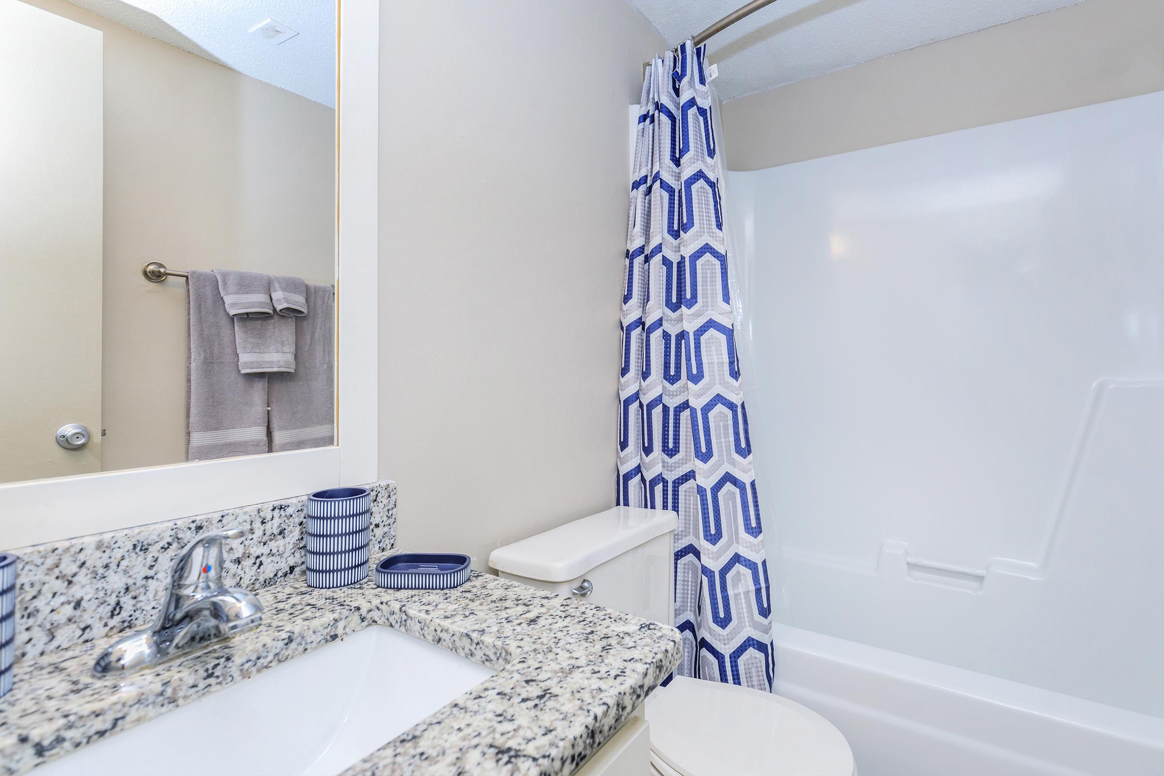 A modern bathroom featuring a white bathtub with a shower curtain that has a geometric blue and white pattern. The sink area includes a speckled granite countertop with a silver faucet and blue decorative items. Soft towels hang on a rod, and light walls create a bright atmosphere.