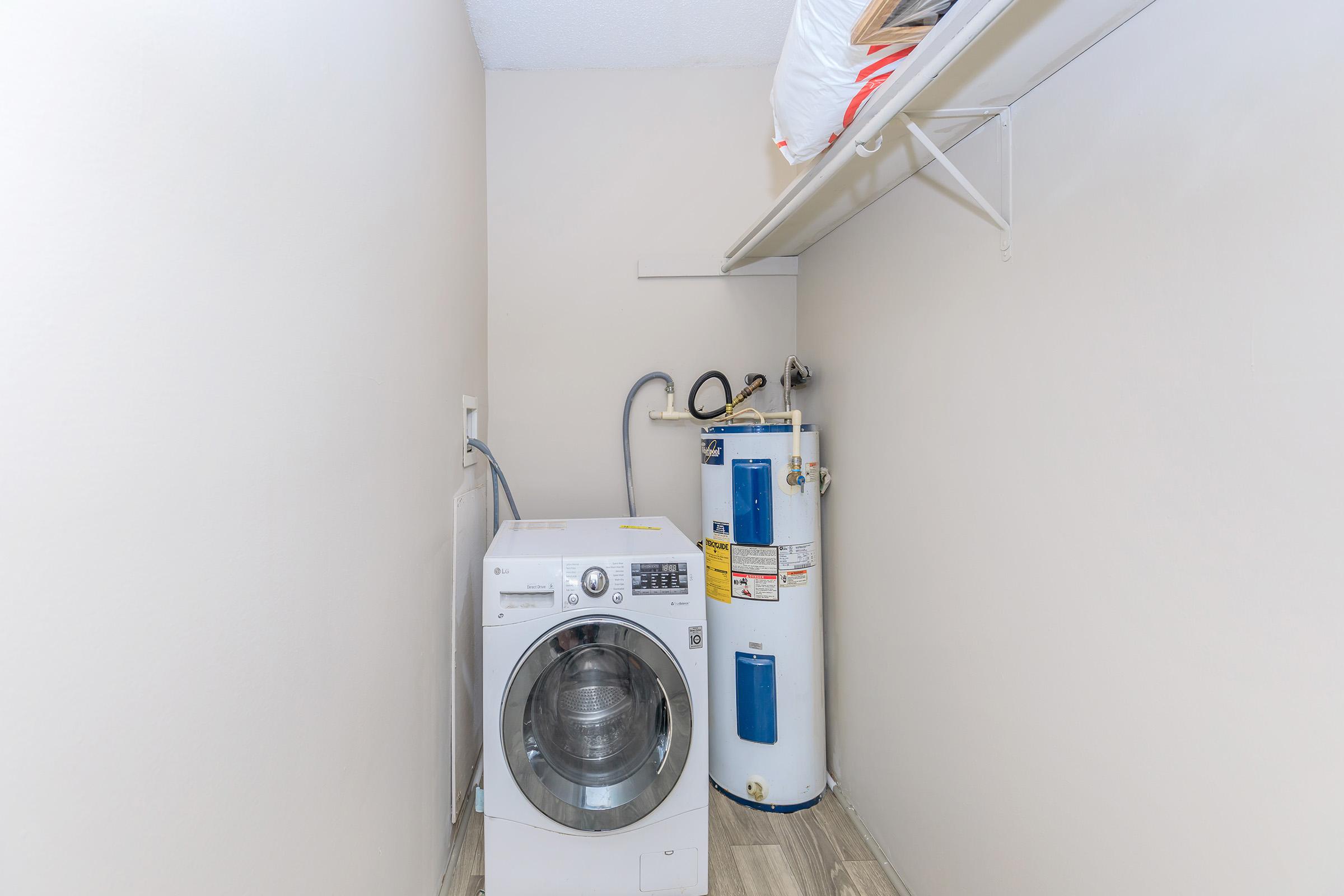 A laundry room featuring a front-loading washing machine next to a water heater. The walls are painted a light neutral color, and there is a shelf above the washer. The flooring is tiled, adding to the clean and functional appearance of the space.