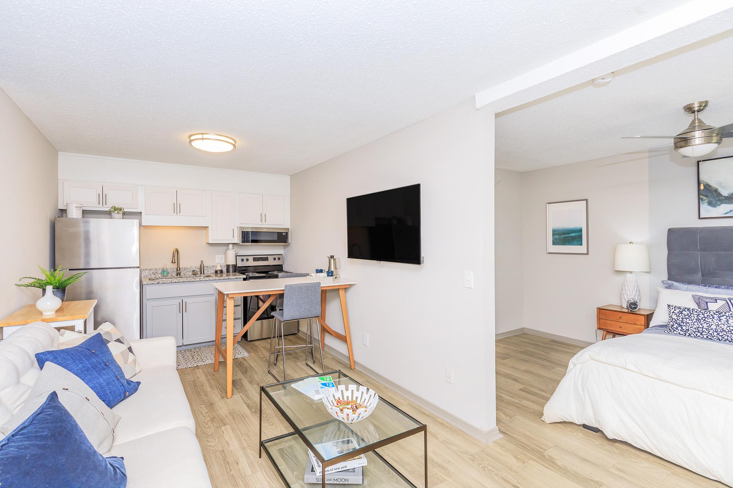 A modern studio apartment with a compact kitchen featuring stainless steel appliances, a small dining table with stools, and a living area with a couch and flat-screen TV. The bedroom area includes a bed with a headboard and a nightstand. Light-colored walls and wood flooring create a bright, inviting atmosphere.