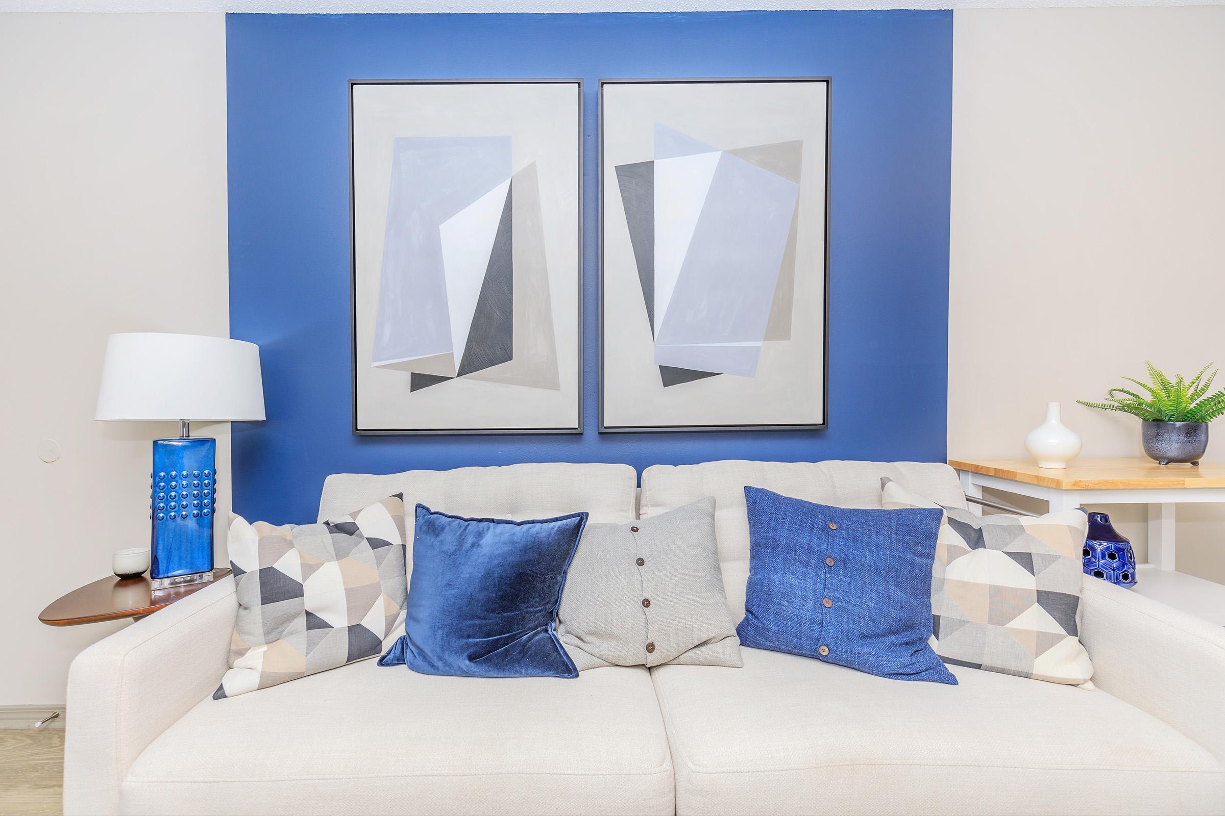 A cozy living room setting featuring a light sofa adorned with various patterned and solid blue cushions. Behind the sofa, there are two abstract framed artworks hanging on a blue accent wall. A stylish lamp and a small plant add to the decor, accompanied by a wooden surface with decorative items.