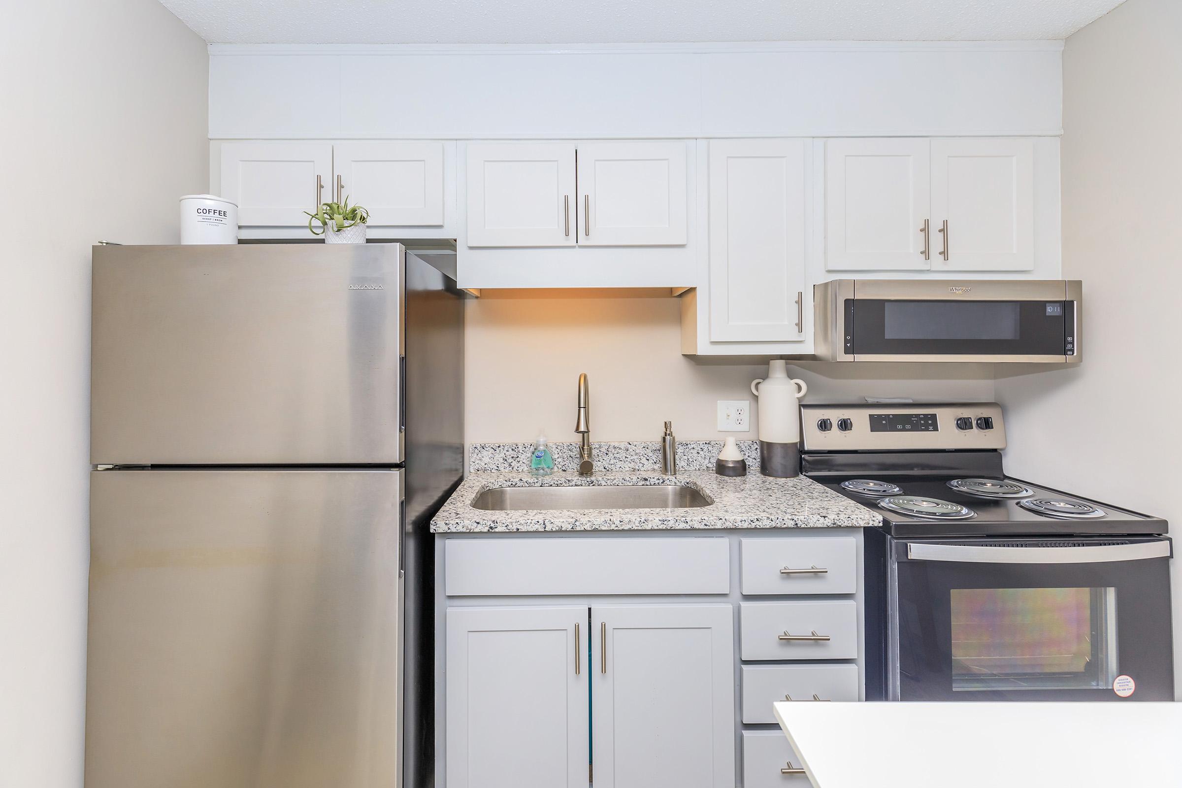 Modern kitchen featuring stainless steel appliances, including a refrigerator, stove, and microwave. The countertop is made of granite, with a sink and various kitchen items displayed. Cabinets above provide storage, and a minimalist aesthetic is evident throughout the space.