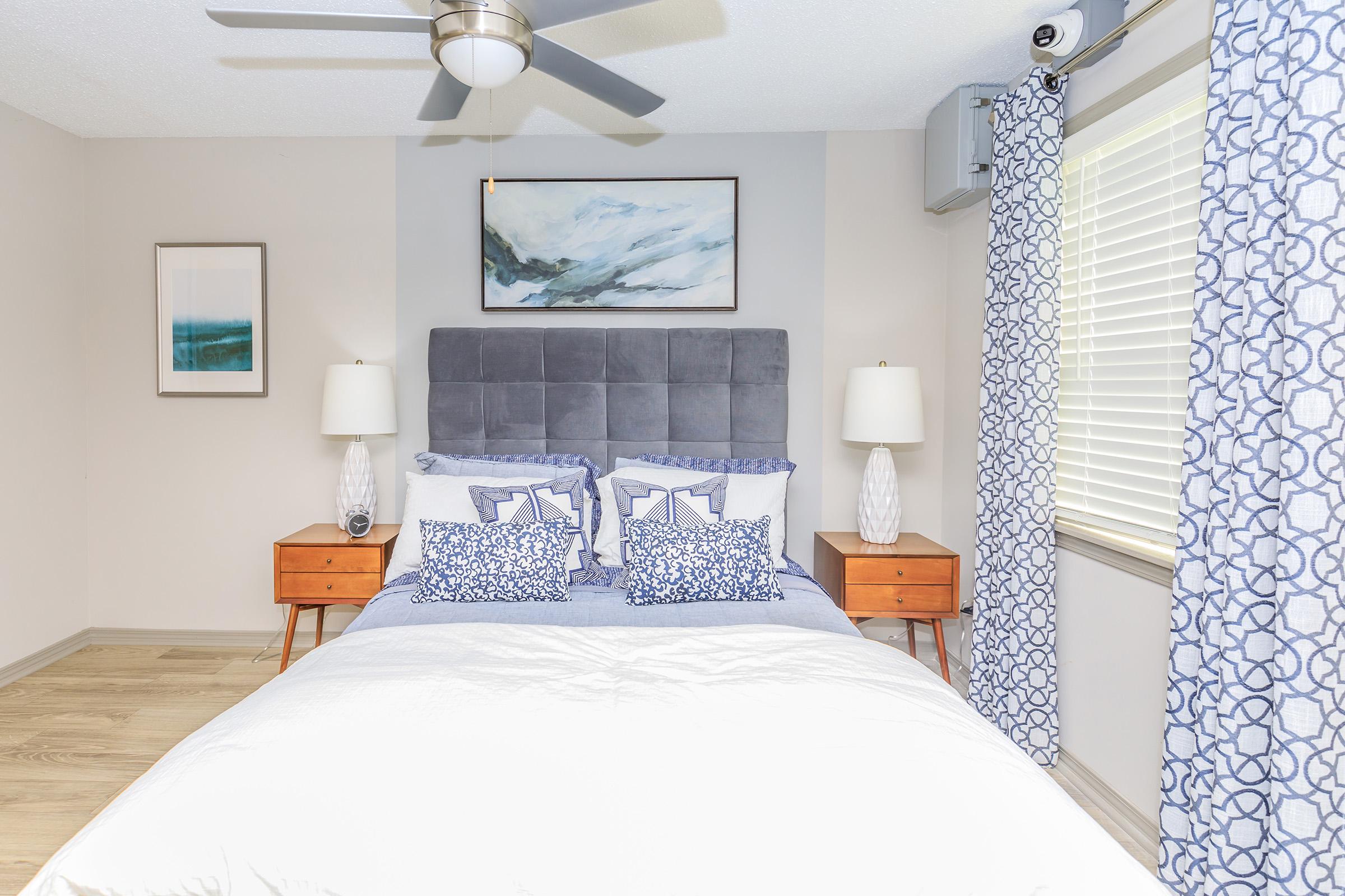 A well-decorated bedroom featuring a gray upholstered headboard with decorative pillows, two wooden nightstands with lamps, and a light-colored comforter. The walls are adorned with a framed artwork, and there are patterned curtains on the windows, creating a cozy atmosphere.