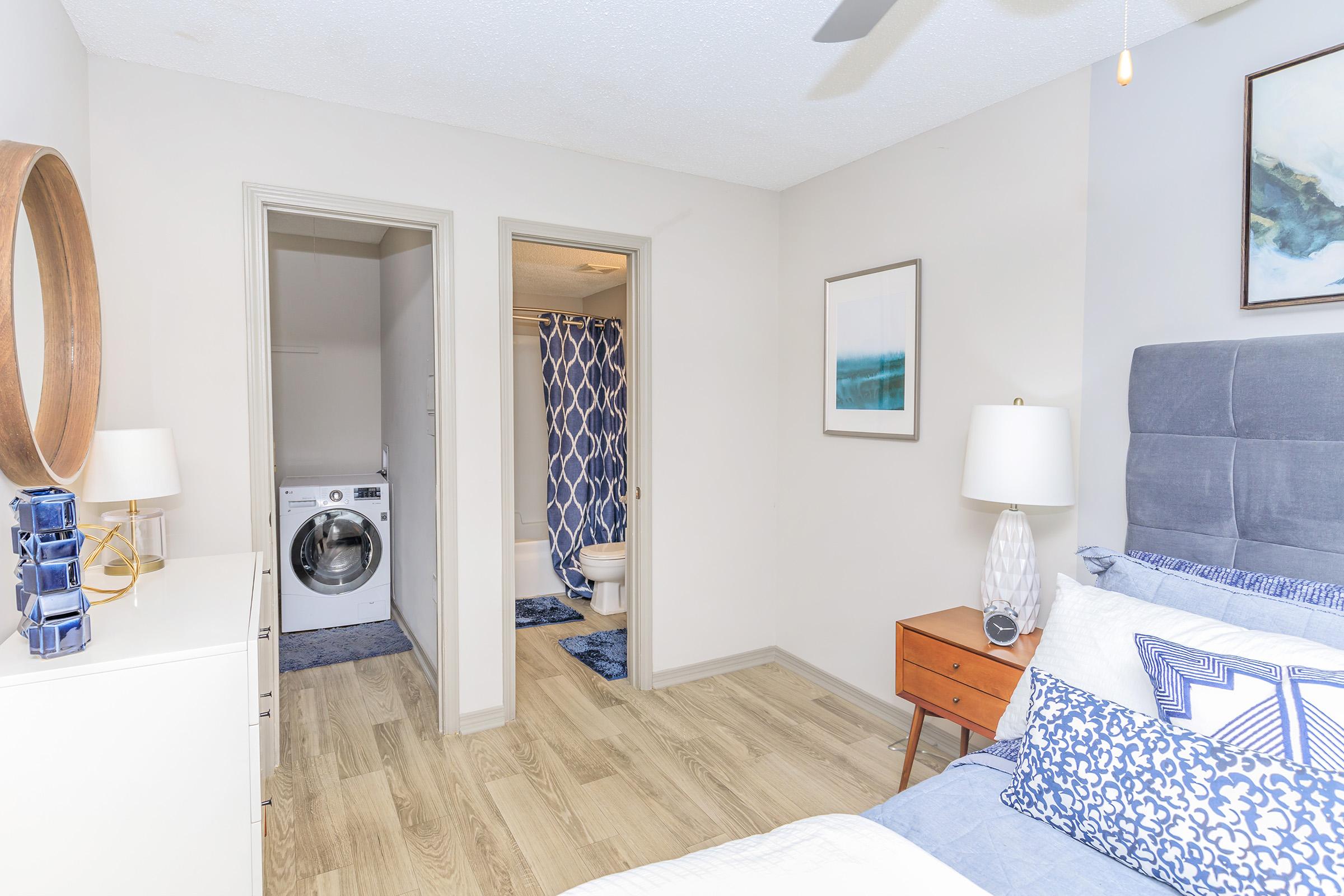 A well-lit bedroom featuring a gray upholstered bed with decorative pillows, a modern nightstand with a lamp, and a circular mirror on the wall. Two doorways lead to a bathroom and laundry area, both with coordinating decor elements. The flooring is light wood, enhancing the spacious feel of the room.