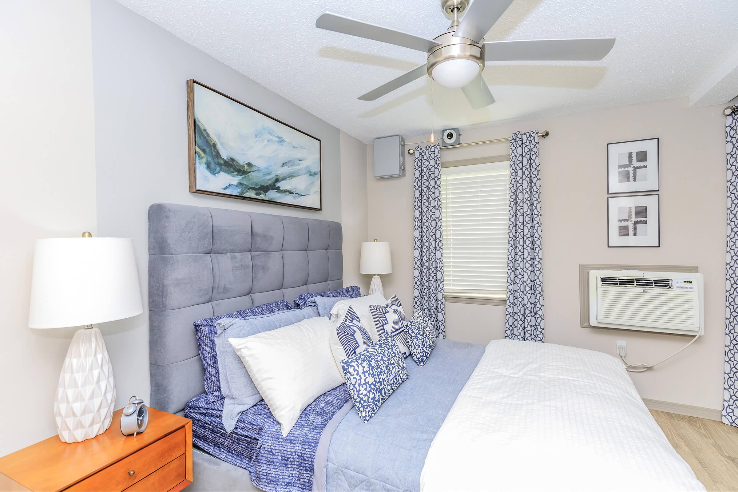A cozy bedroom featuring a grey upholstered headboard with decorative pillows, a blue-and-white color scheme, a wooden nightstand with a lamp, framed artwork on the wall, and a window with patterned curtains. An air conditioning unit is visible beside the window, adding comfort to the space.