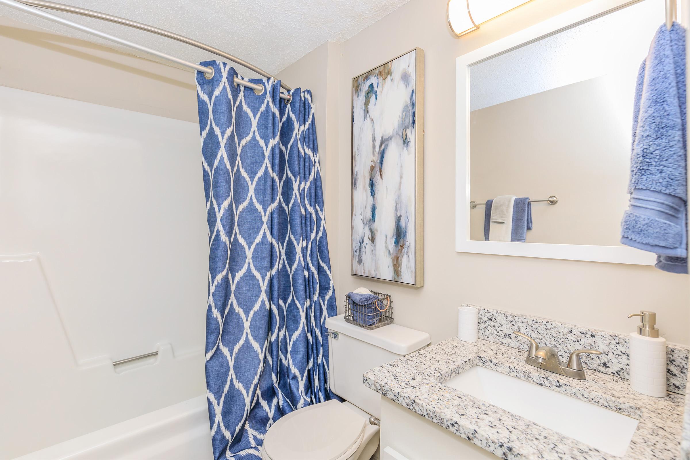 A clean, modern bathroom featuring a white bathtub with a shower curtain in a blue geometric pattern, a granite countertop with a sink, a small storage container, a wall-mounted mirror, and a painting. Soft blue towels hang nearby, enhancing the aesthetic appeal.