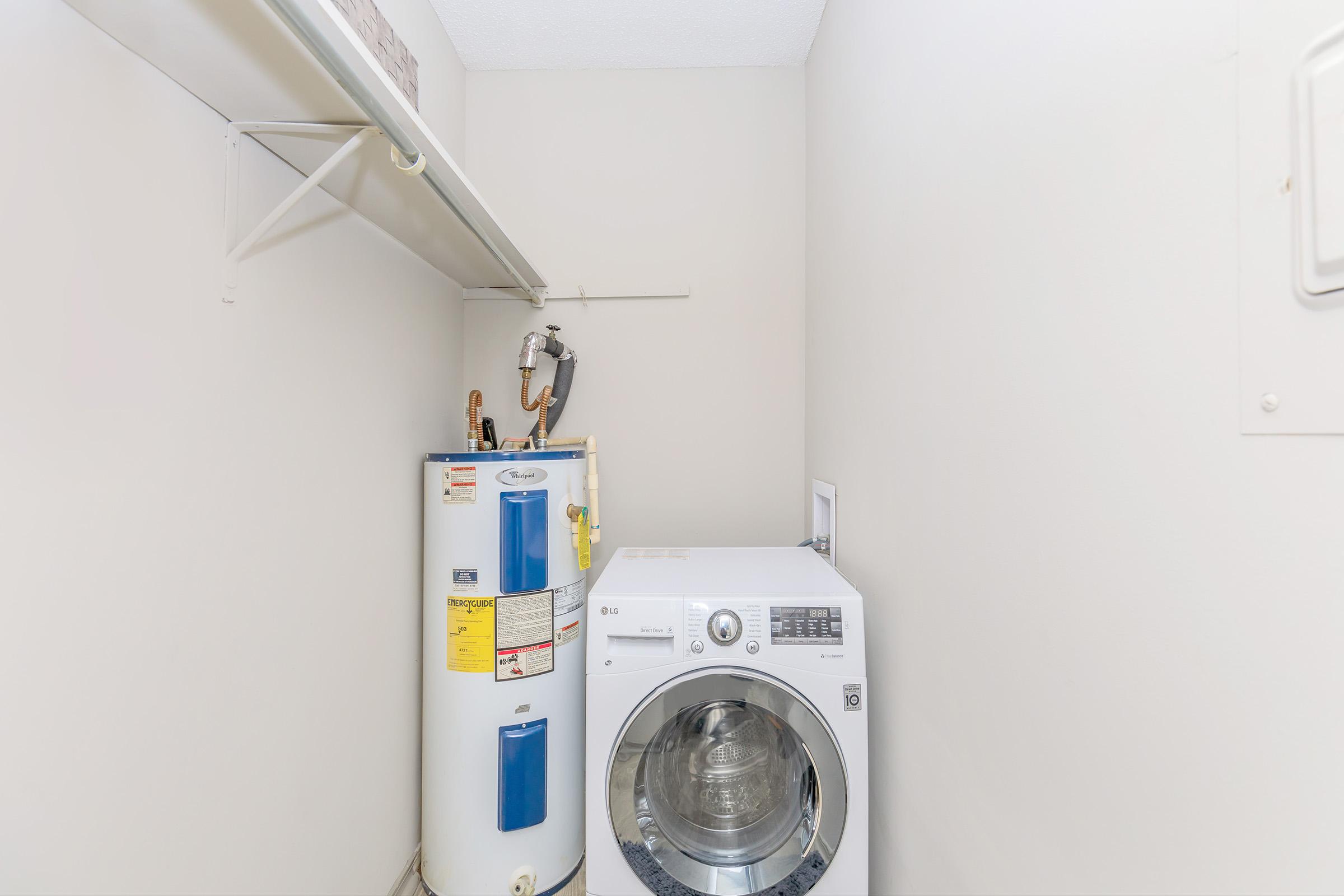A small laundry room featuring a white washing machine next to a water heater. The room is painted light gray with a shelf above the washer. A small vent is visible on the wall.