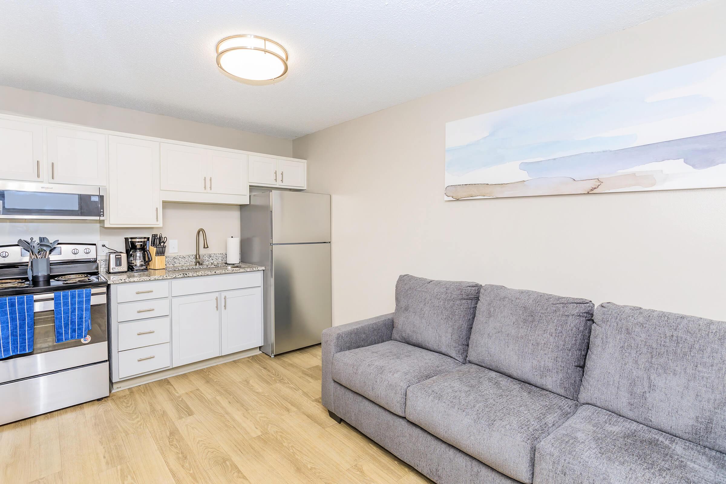 Modern kitchen featuring stainless steel appliances, including a stove and refrigerator, with a granite countertop and white cabinetry. A cozy gray sofa is positioned in the living area, complemented by a wall art piece depicting a serene landscape. The flooring is light wood.