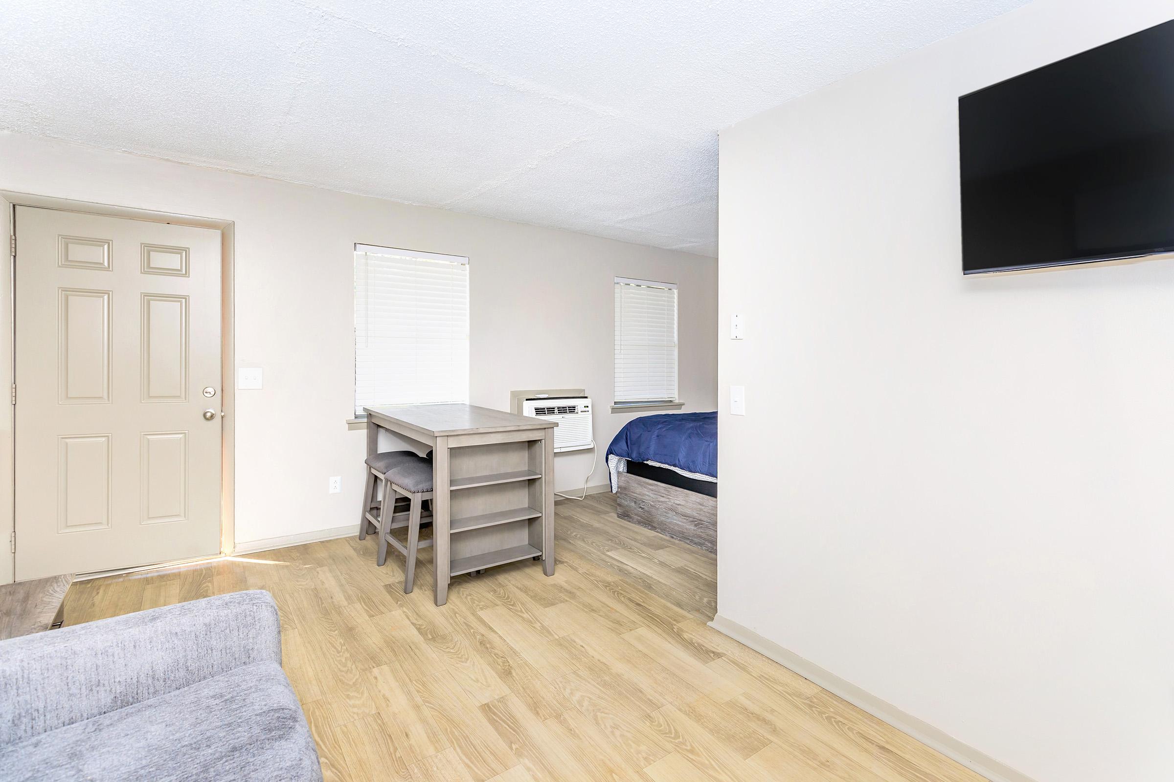 A light-colored interior room featuring a gray sofa, a wooden dining table with chairs, a bed with blue bedding, a window with blinds, an air conditioning unit, and a wall-mounted television. The flooring is light wood, and there is a door leading to another area.