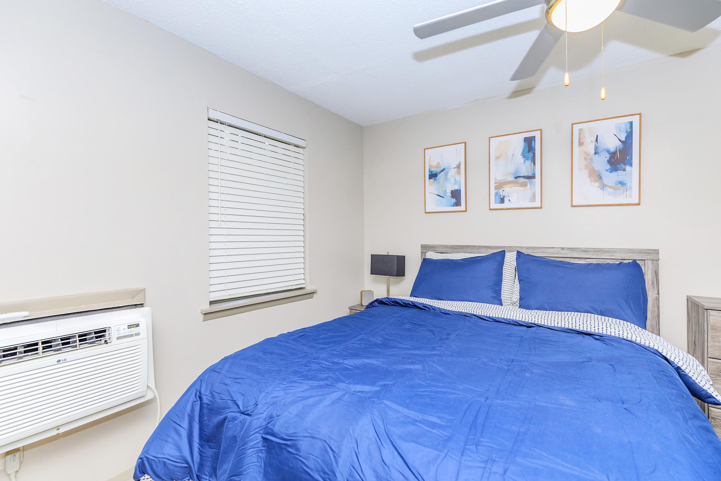 A cozy bedroom featuring a large bed with blue bedding, a ceiling fan, and an air conditioning unit on the wall. The room has light-colored walls and three abstract framed pictures above the bed, with a window adorned by white blinds allowing natural light to enter.