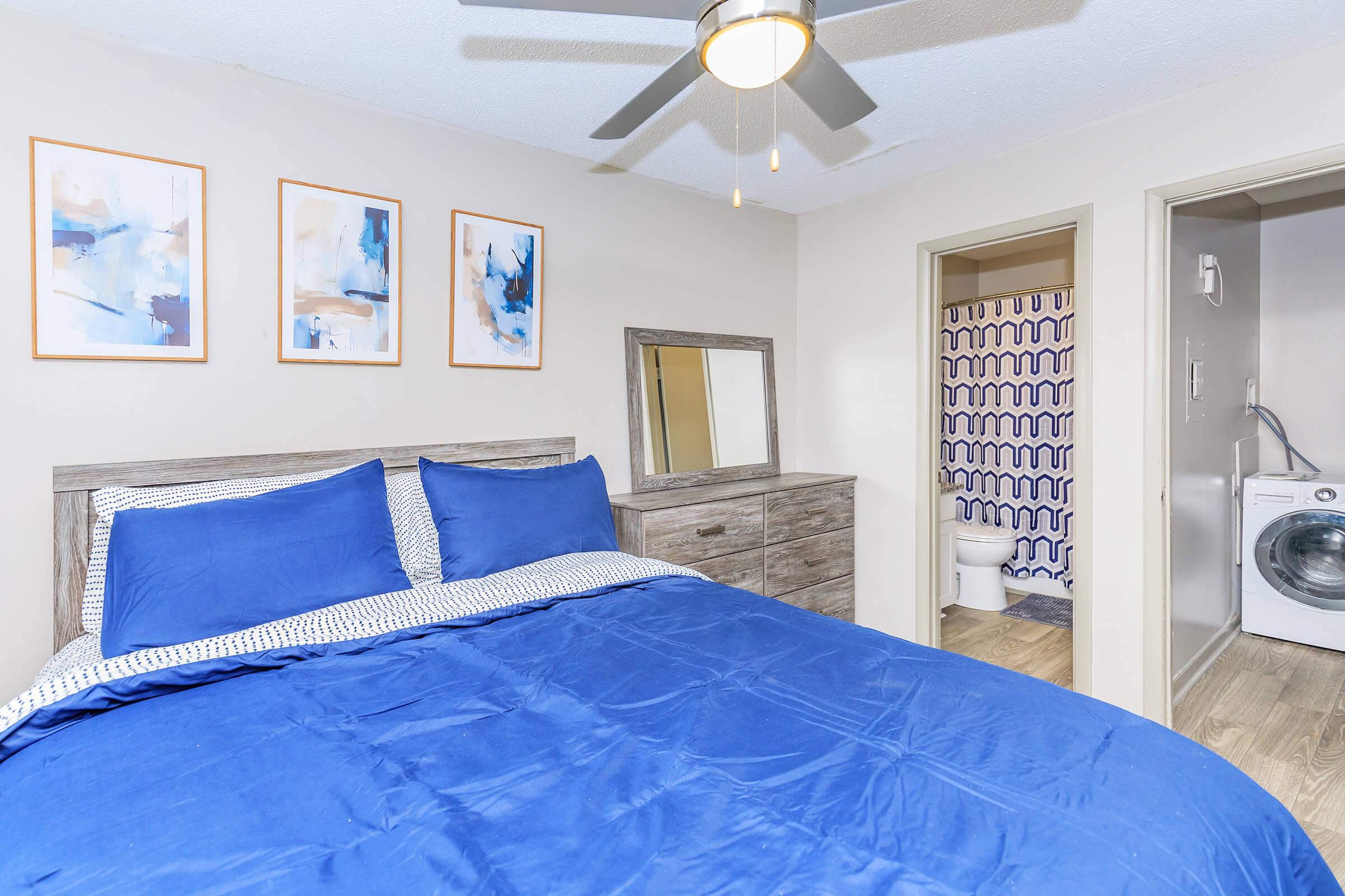 A cozy bedroom featuring a large bed with a blue comforter and matching pillows. The room has a dresser and three framed abstract art pieces on the wall. A doorway leads to a bathroom area, which includes a visible toilet. A washing machine is also present in the corner of the room.