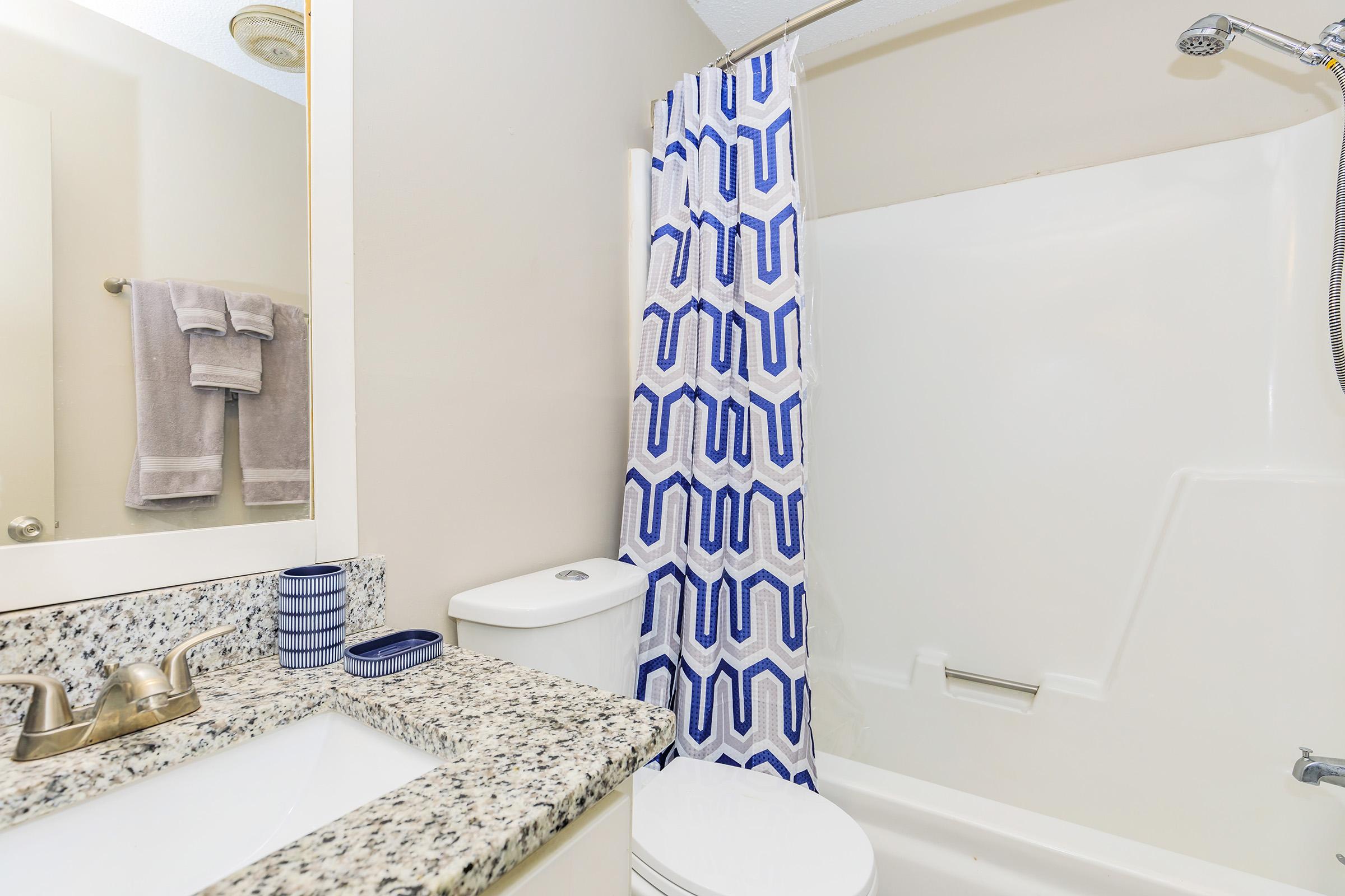 A well-lit bathroom featuring a white bathtub with a decorative shower curtain in blue and white geometric patterns. The sink area has a granite countertop, with a modern faucet, a small decorative container, and a towel rack with gray towels. The walls are a neutral color, enhancing the clean aesthetic.