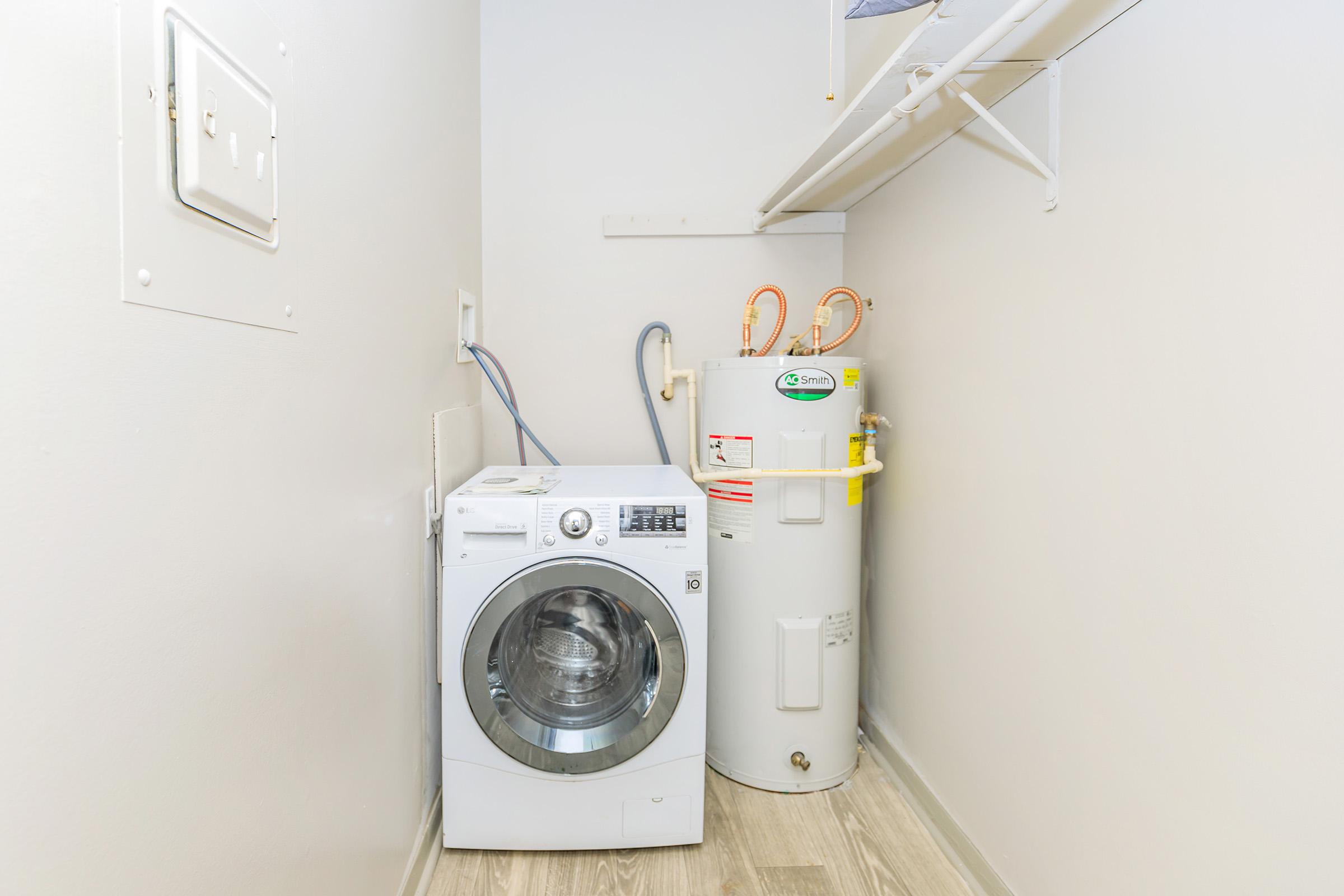 A laundry room featuring a white washing machine next to a cylindrical water heater. The walls are painted a light color, and the floor has a wood-like appearance. A shelf is mounted above the appliances, providing storage space.