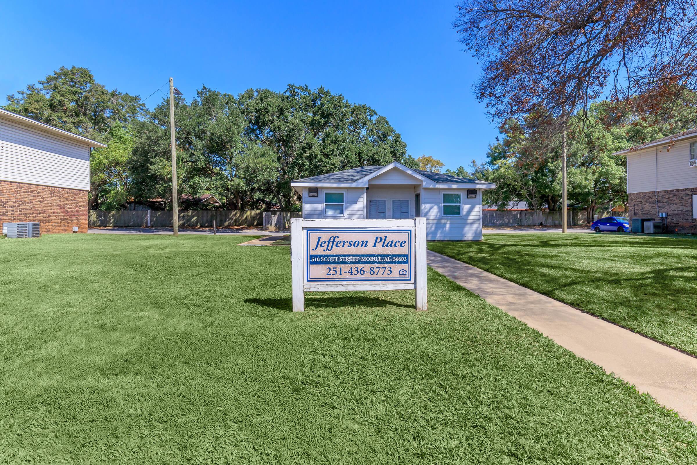 A small single-story house with a sign in front reading "Jefferson Place" and contact information. The house is flanked by trees and has a well-maintained lawn. A paved walkway leads to the entrance. In the background, there are other residential buildings.