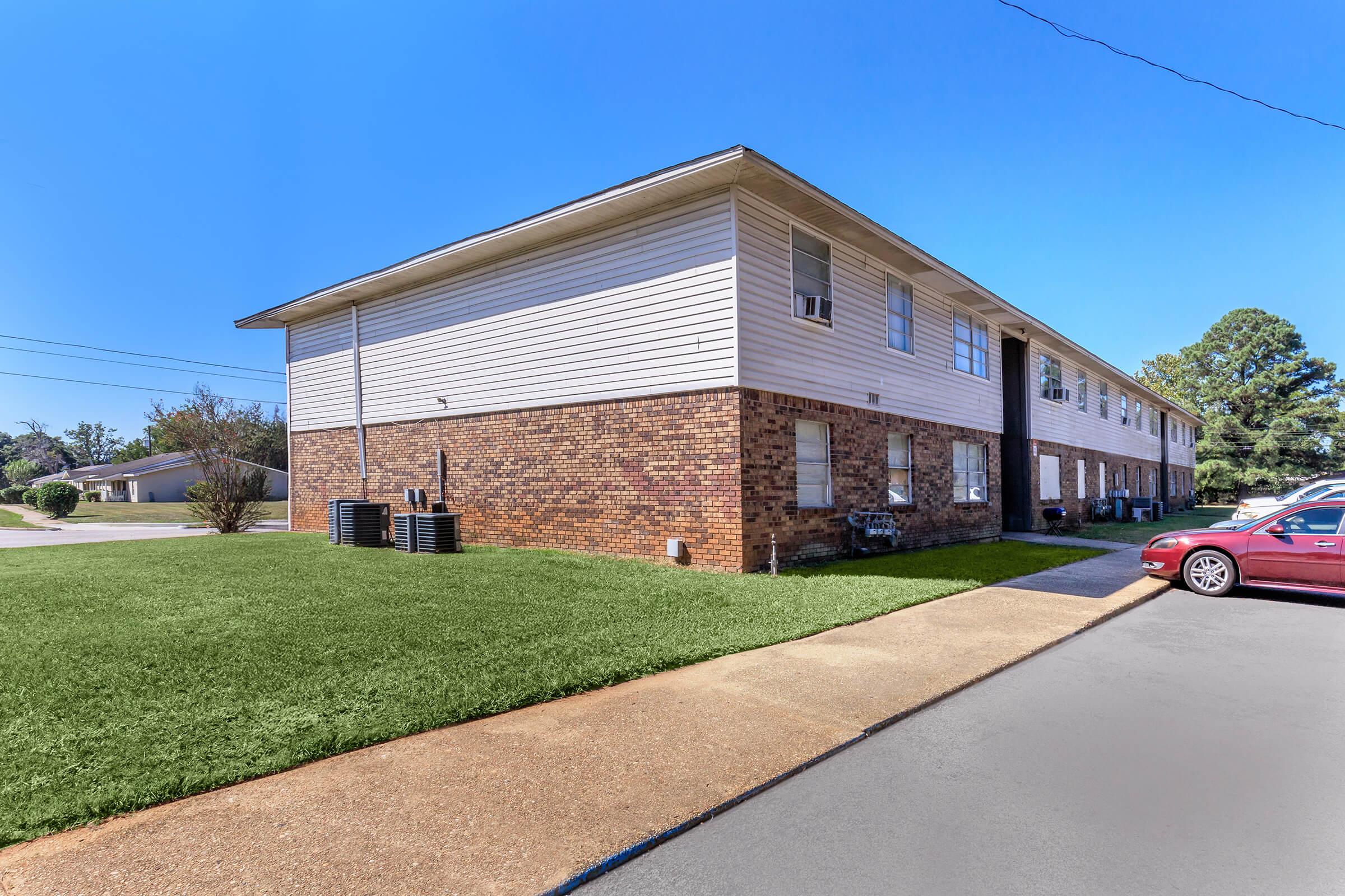 Side view of a two-story apartment building with a brick and white exterior. The lawn in front is well-maintained, and there are air conditioning units visible. A paved pathway leads from the parking area to the entrance of the building, which has several windows. The sky is clear and blue.