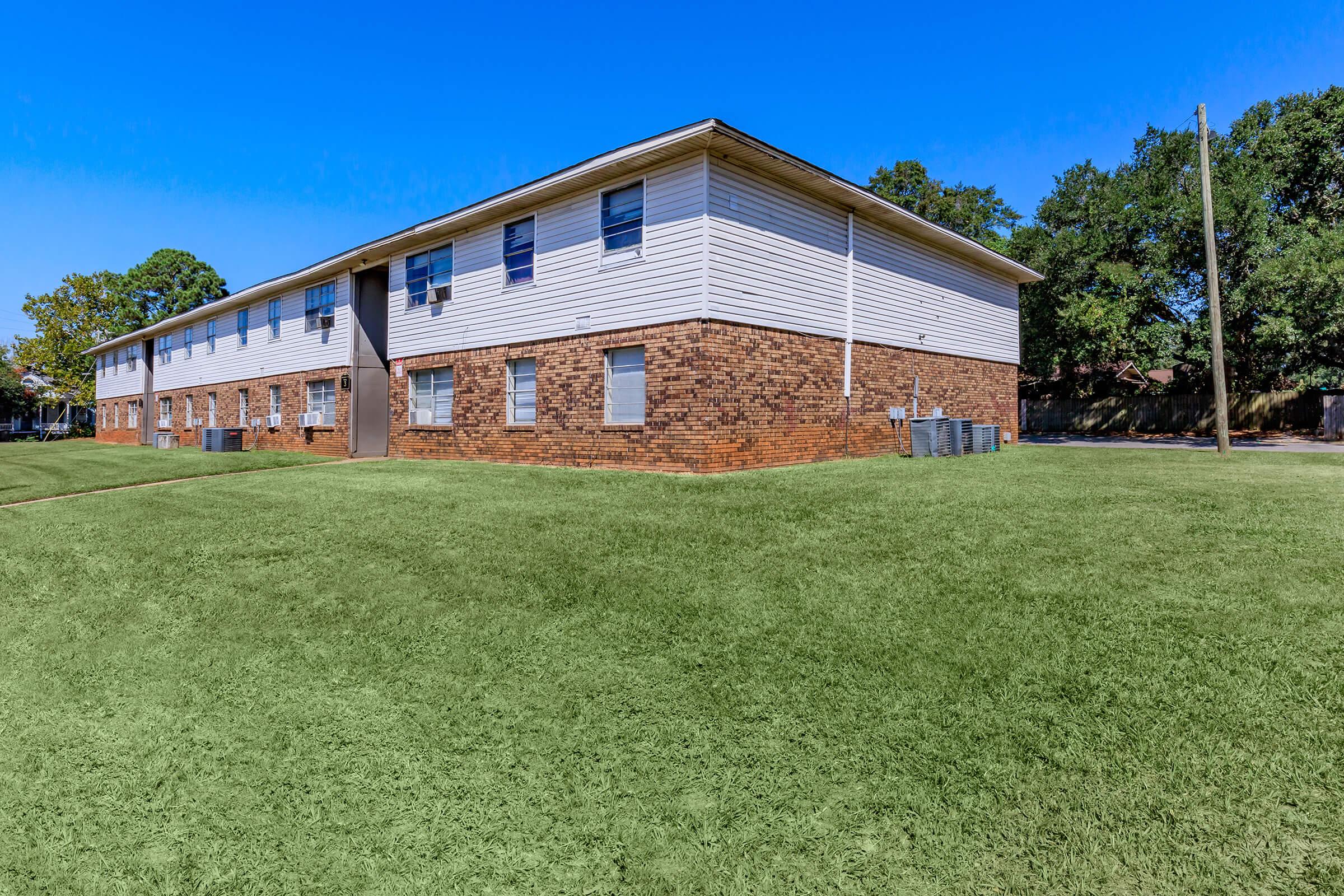 A large two-story apartment building with a mix of brick and white siding. The building features multiple windows and a central entrance, surrounded by a well-maintained green lawn under clear blue skies. Air conditioning units are visible outside the building.