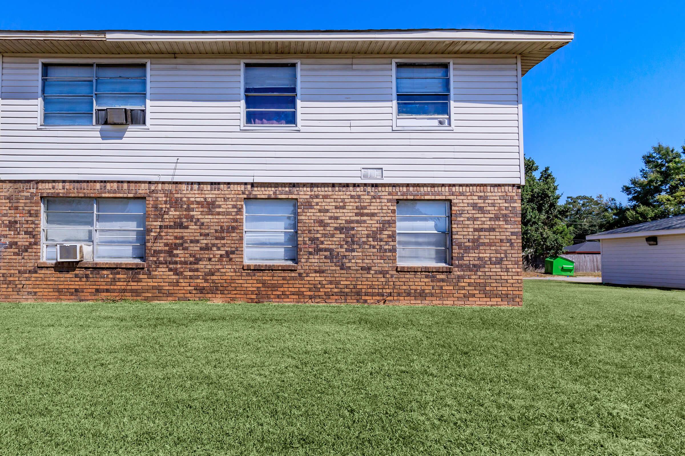 A two-story building with a brick and siding exterior. The ground level features three windows, two of which are framed in brick, and an air conditioning unit. The grassy area in front is well-maintained, and there is a separate building visible in the background. The sky is clear and blue.