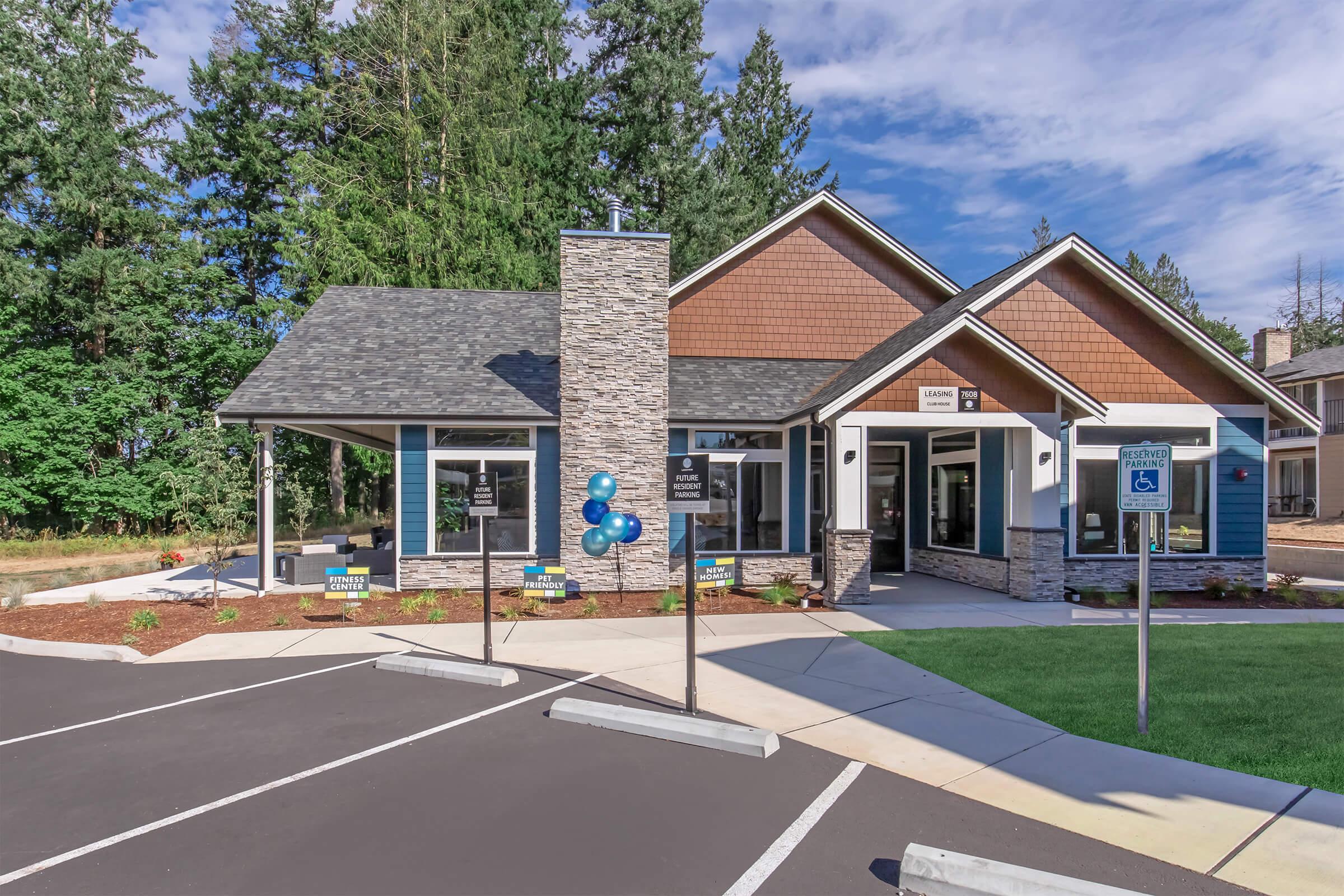 A modern single-story building with a stone and wood facade, surrounded by greenery. The entrance features large windows and a welcoming porch. Several signs are placed in front, indicating various services, and blue balloons are tied to a post, adding to the inviting atmosphere.