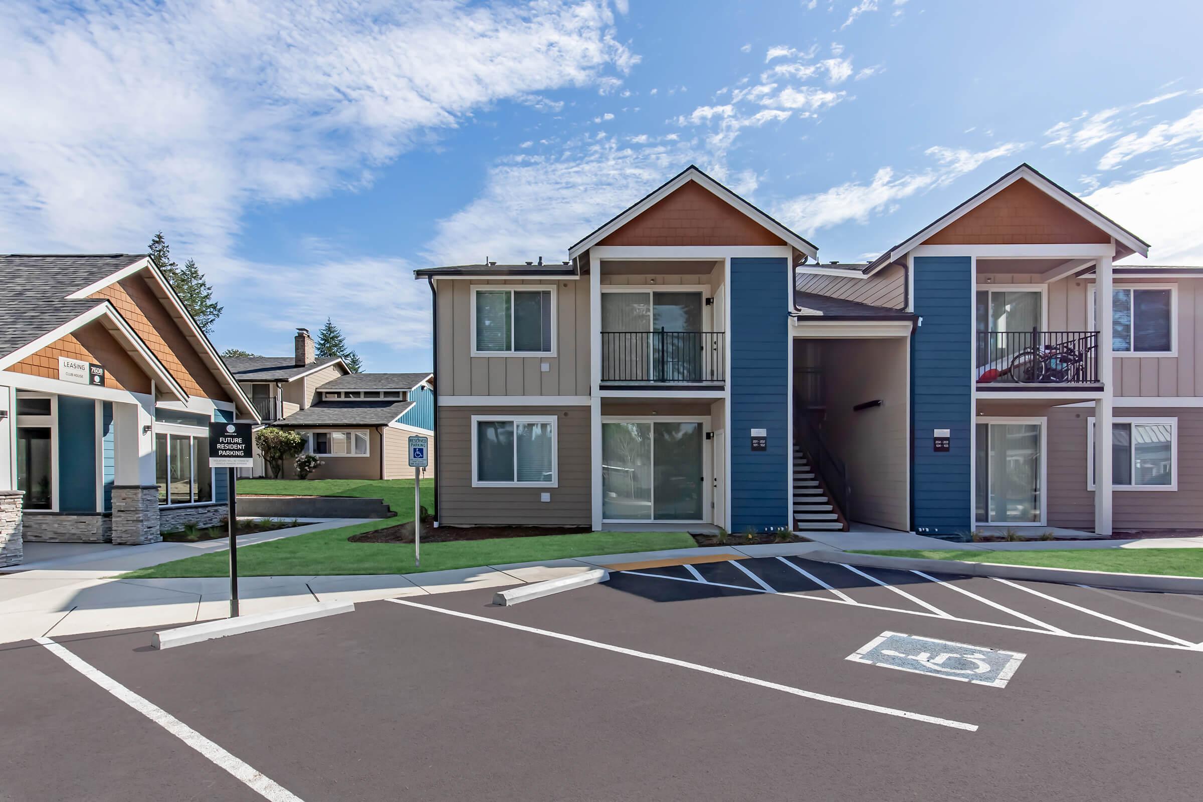 Two-story apartment building featuring a mix of tan and blue siding with balconies. A sidewalk leads up to the entrance, and a designated handicapped parking space is visible in the foreground. The sky is clear with a few clouds, and the surrounding area includes landscaped grass and additional buildings.