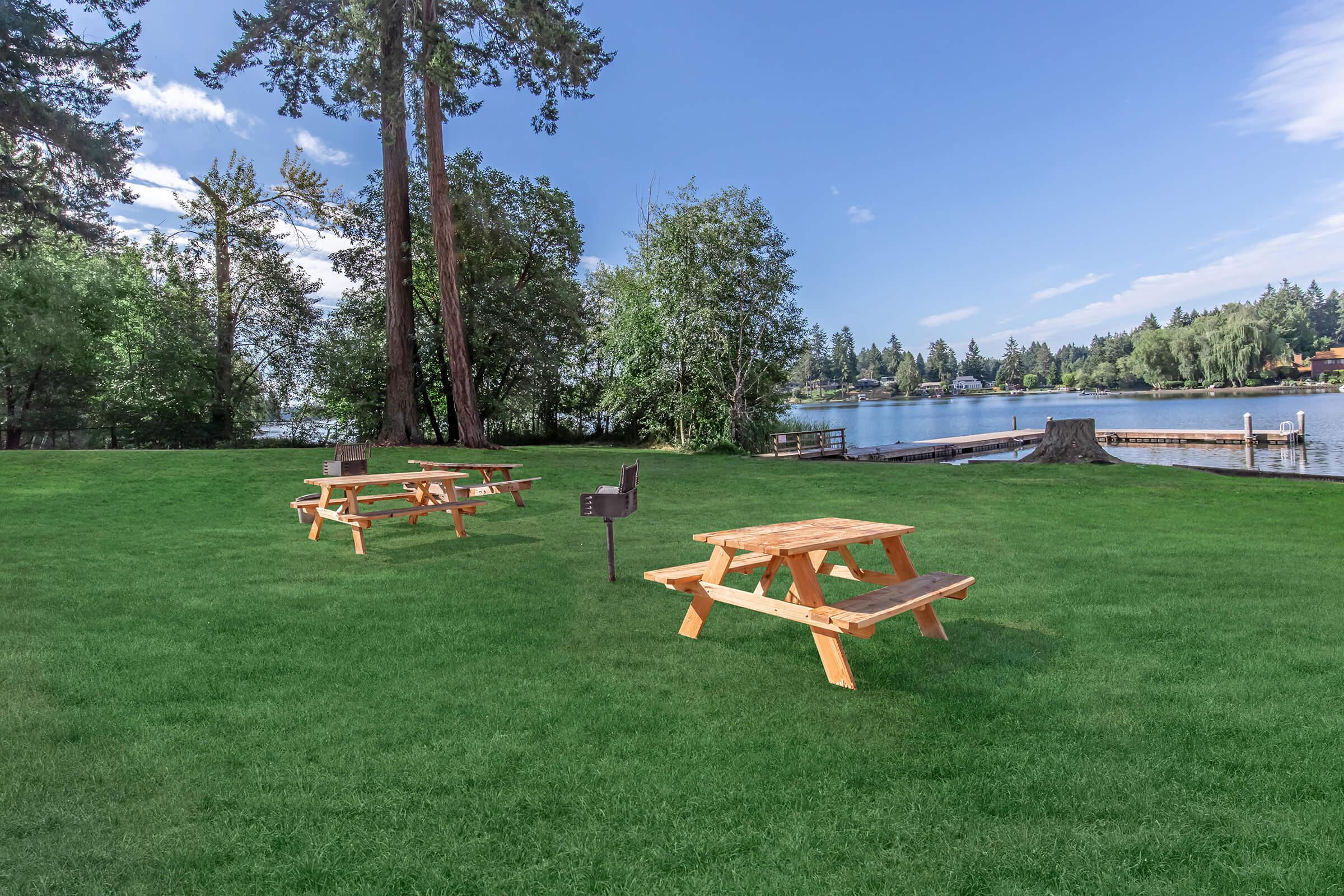 A serene view of a lakeside picnic area featuring several wooden picnic tables on a lush green lawn. In the background, trees frame the lake and a dock extends into the water under a clear blue sky with scattered clouds. Ideal for outdoor gatherings and relaxation.
