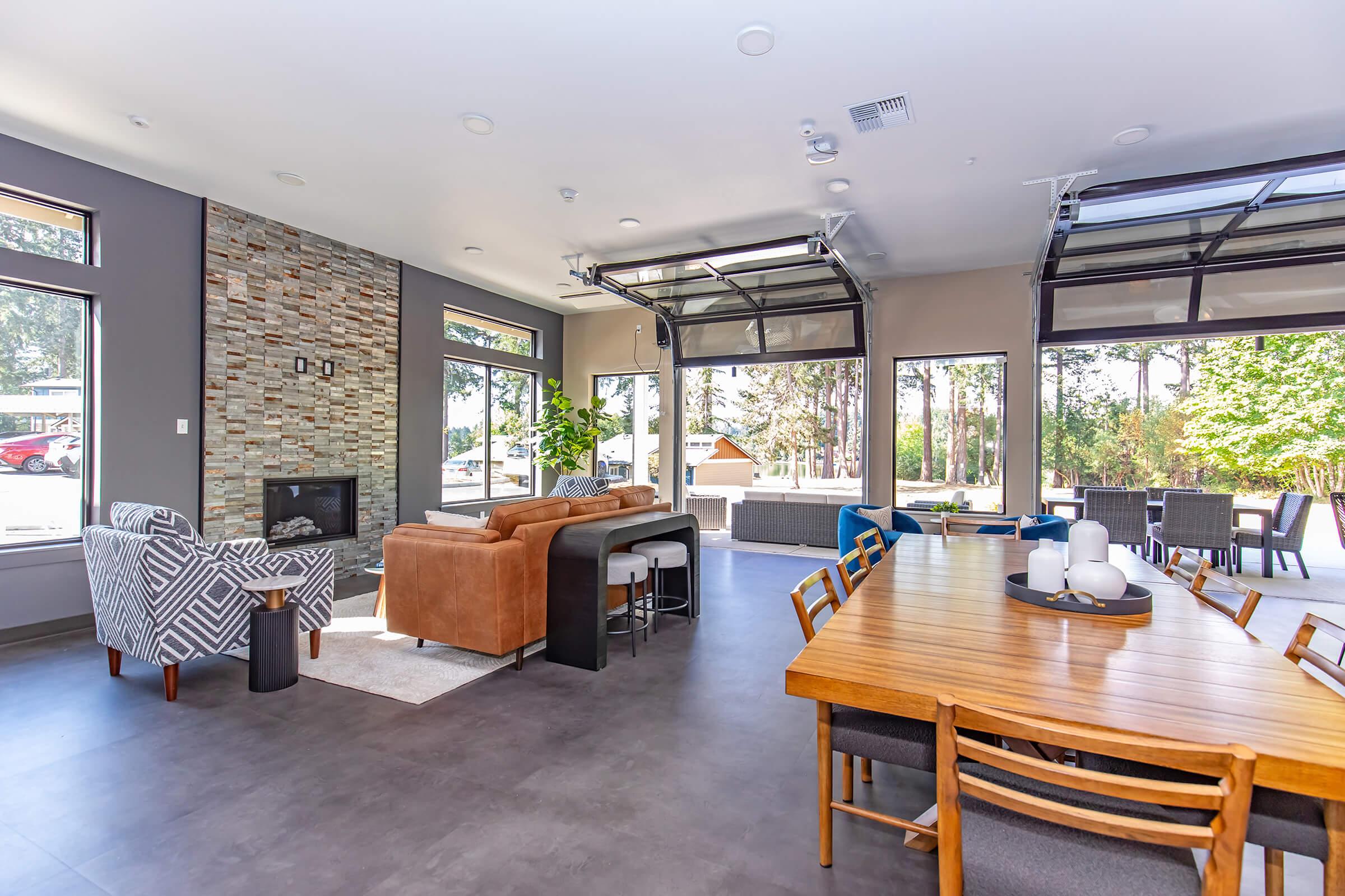 Modern living space featuring a cozy seating area with a sofa and armchair, a dining table with chairs, large windows, and a stone accent wall. The room is well-lit and opens up to a patio with outdoor seating, surrounded by trees. The decor is stylish and contemporary, creating a welcoming atmosphere.