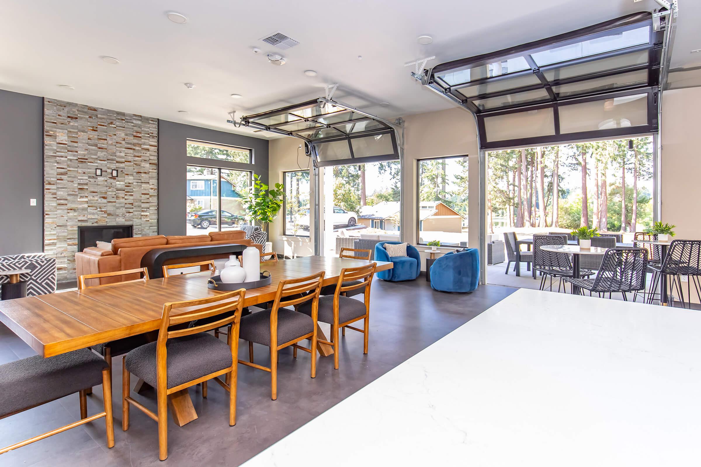 Modern open-concept living space featuring a dining area with a wooden table and chairs, a cozy seating arrangement with a sofa, colorful accent chairs, and large windows allowing natural light. A stone wall adds texture, while a patio area is visible outside, surrounded by trees.