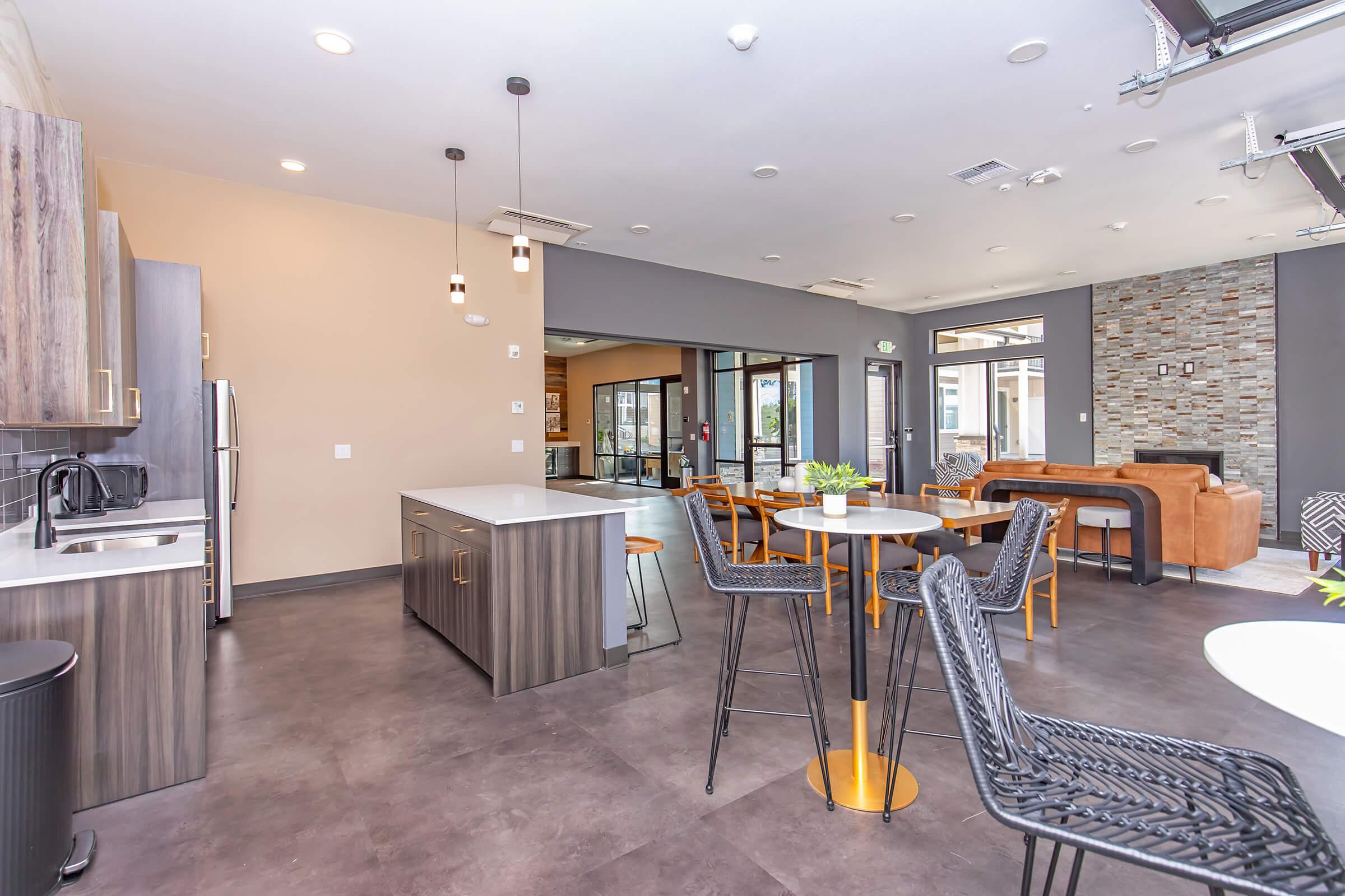 Modern interior of a spacious communal kitchen and lounge area. Features sleek cabinetry, stainless steel appliances, and a variety of seating options including tables and chairs. Large windows allow natural light, with a cozy seating area visible in the background. Stylish decor adds to the inviting atmosphere.